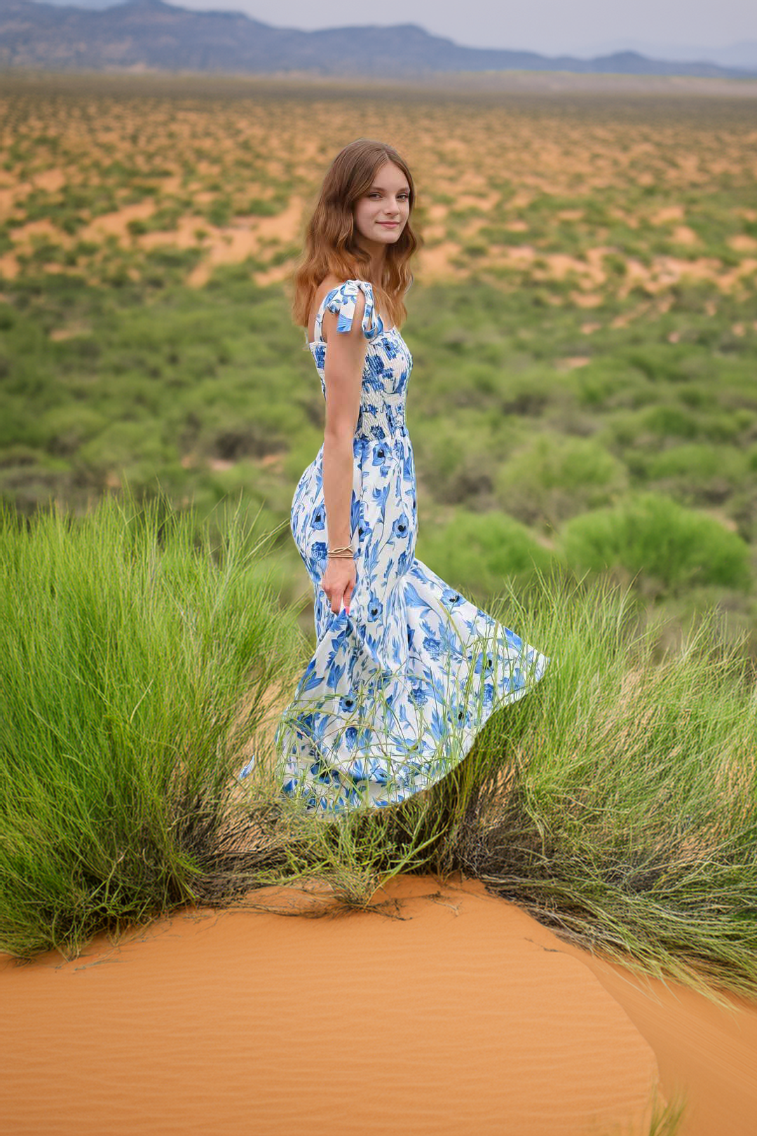 A figure in a flowing blue and white floral dress stands gracefully atop orange sand dunes with desert brush in the background.