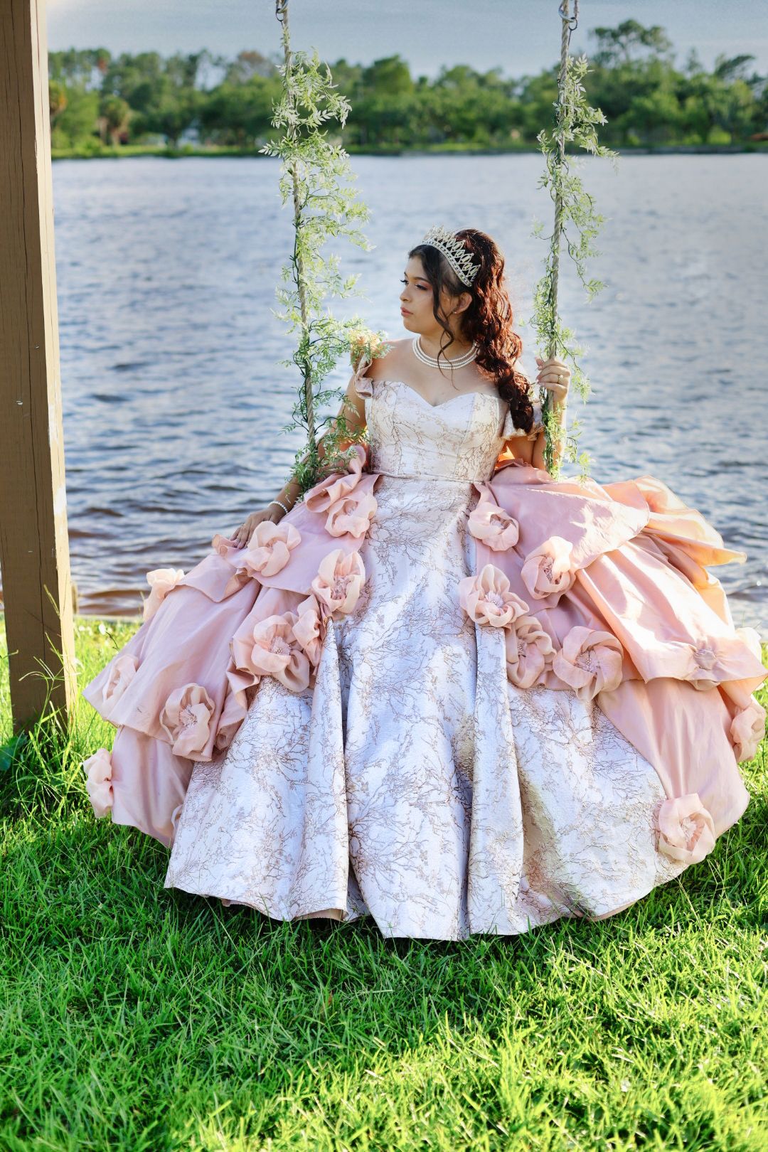 Young woman in elegant pink quinceañera ball gown sitting on a floral swing near a serene lake.