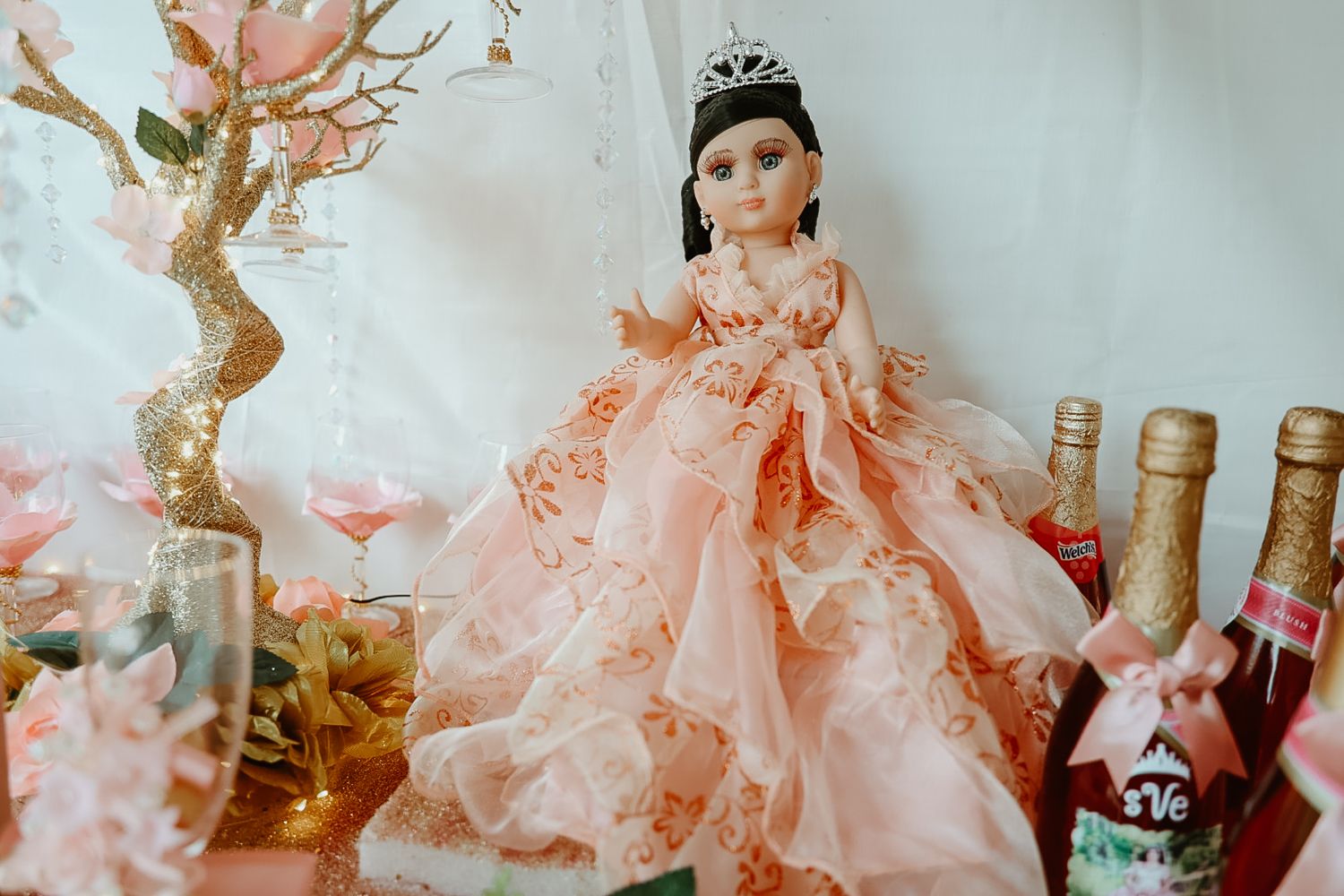 Elegant quinceañera doll in peach gown with tiara displayed on table with champagne bottles.