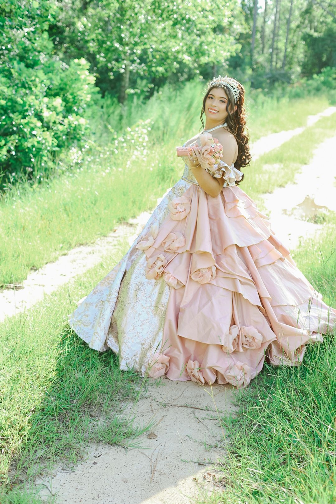 Young woman in a layered pink ball gown with floral details poses on a garden path surrounded by lush greenery.