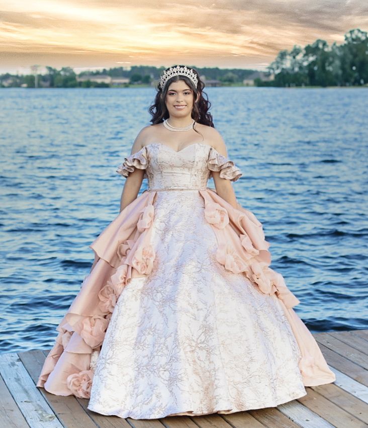 Young woman in elegant blush quinceañera ball gown and tiara poses on a dock with a serene lake at sunset.