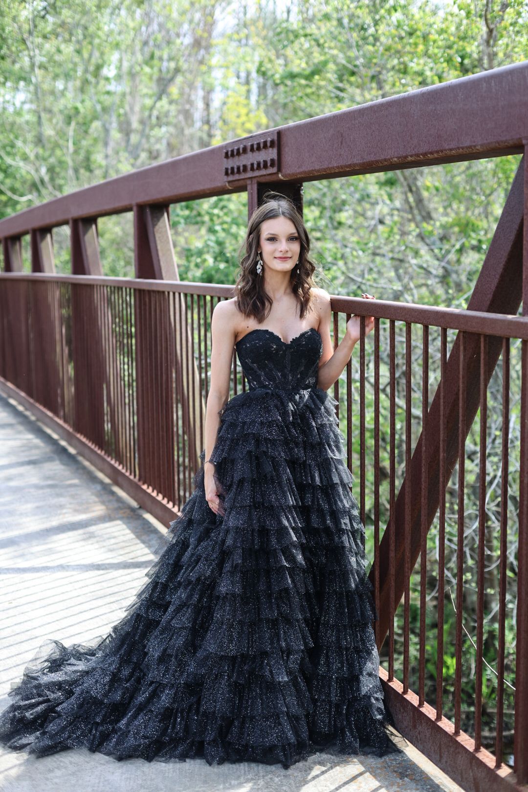 Young woman in elegant black strapless ruffled ball gown posing on a rustic red metal bridge outdoors.