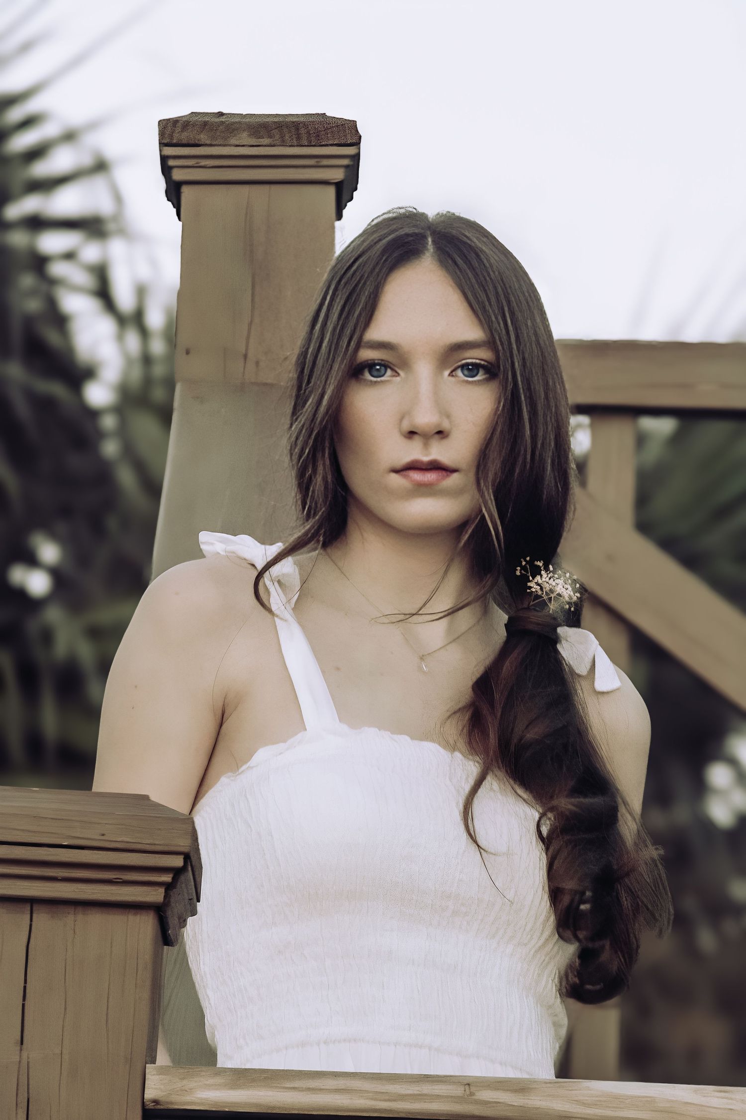 Young woman with long dark hair and floral accessory poses in white smocked top near wooden railing outdoors.