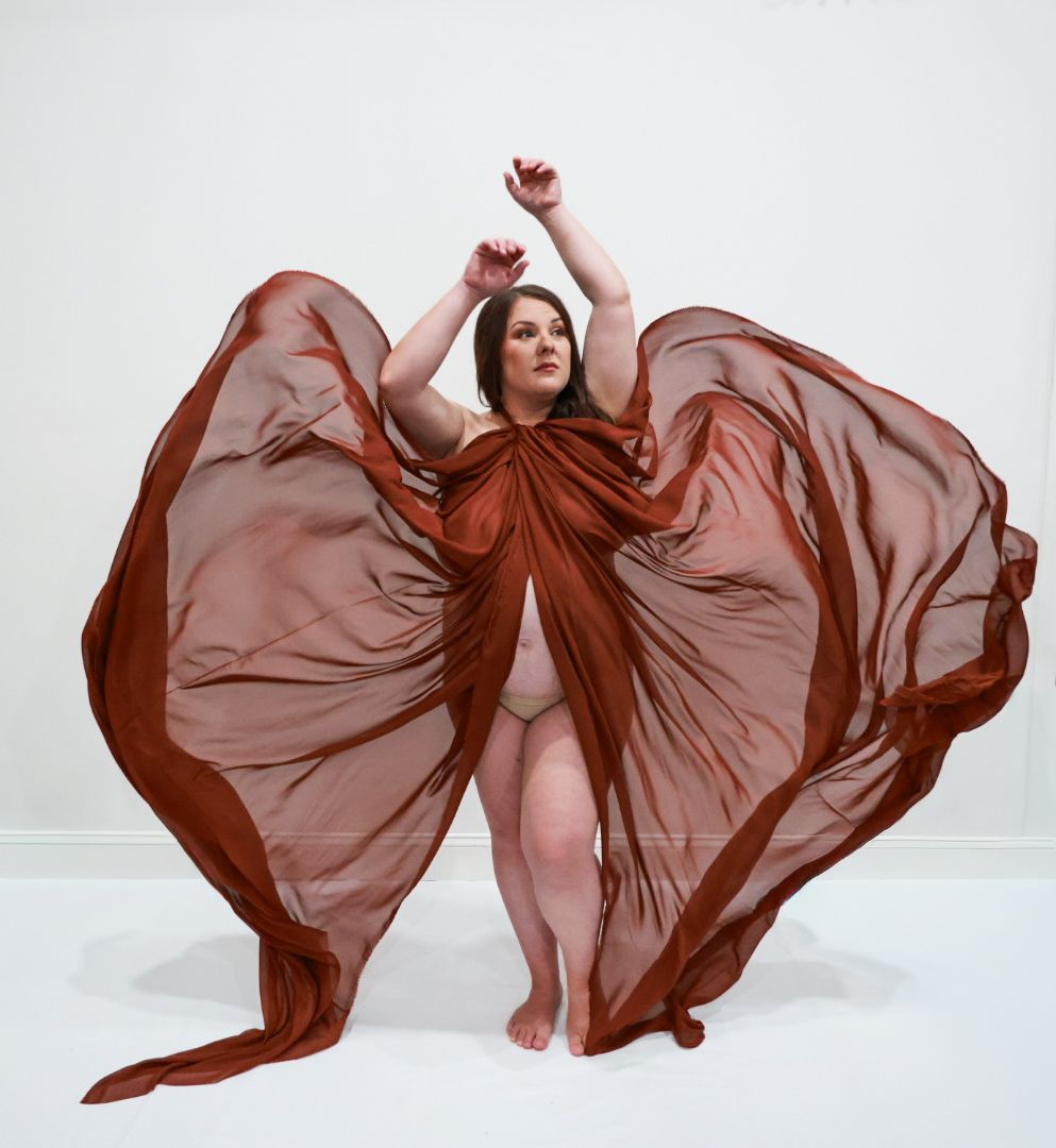 A plus-size woman poses gracefully draped in flowing rust-colored sheer fabric against a white background.