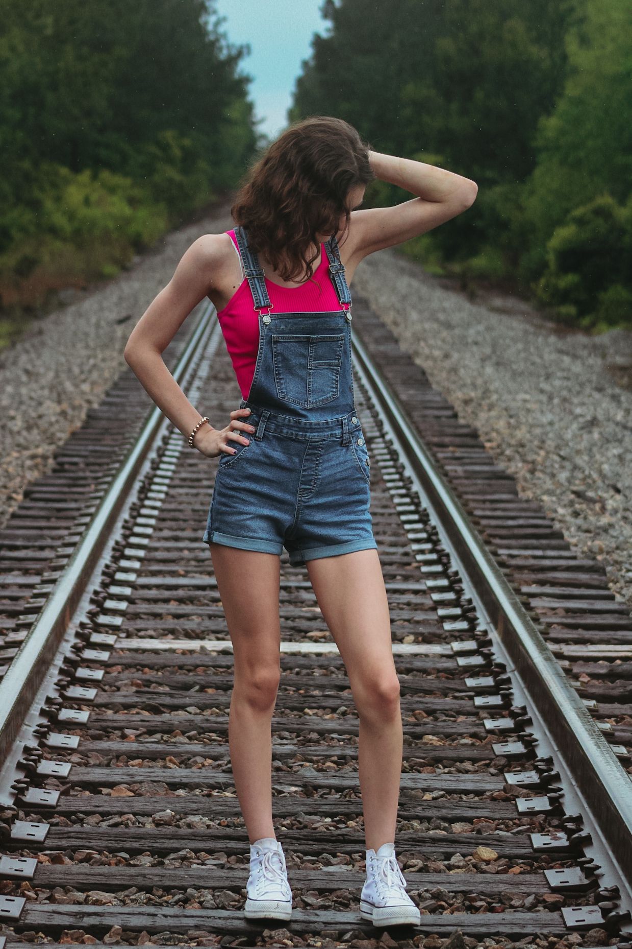 Young woman in denim overalls and pink top posing on railroad tracks surrounded by lush green trees.