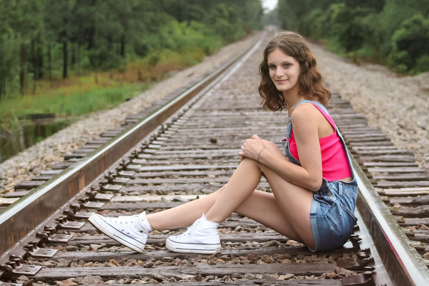 Young woman in pink top and denim shorts sitting on railroad tracks surrounded by lush green forest.