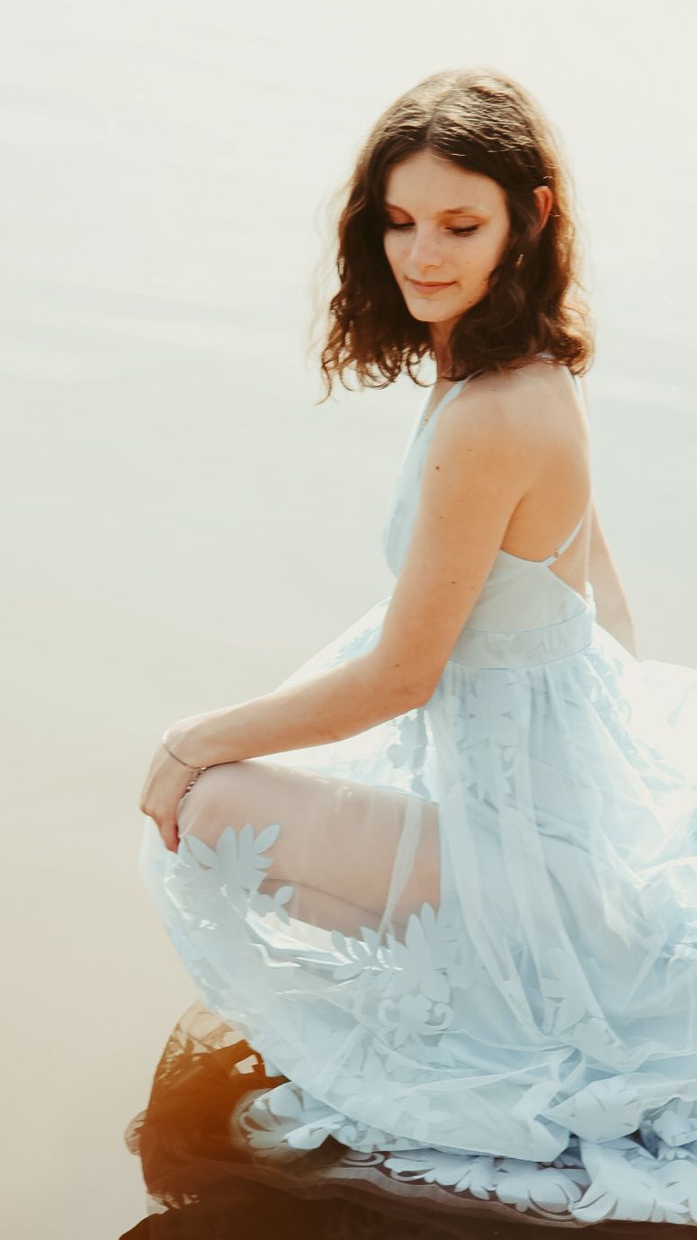 Woman in light blue floral lace dress sitting gracefully near water, looking down in a serene outdoor setting.