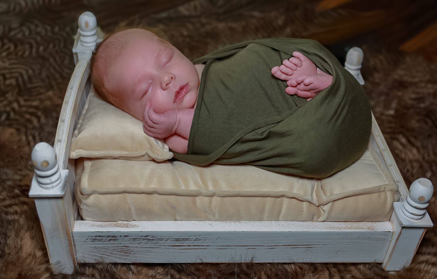 A peaceful newborn wrapped in olive green fabric sleeps soundly on cream bedding in an antique-style white bed frame.