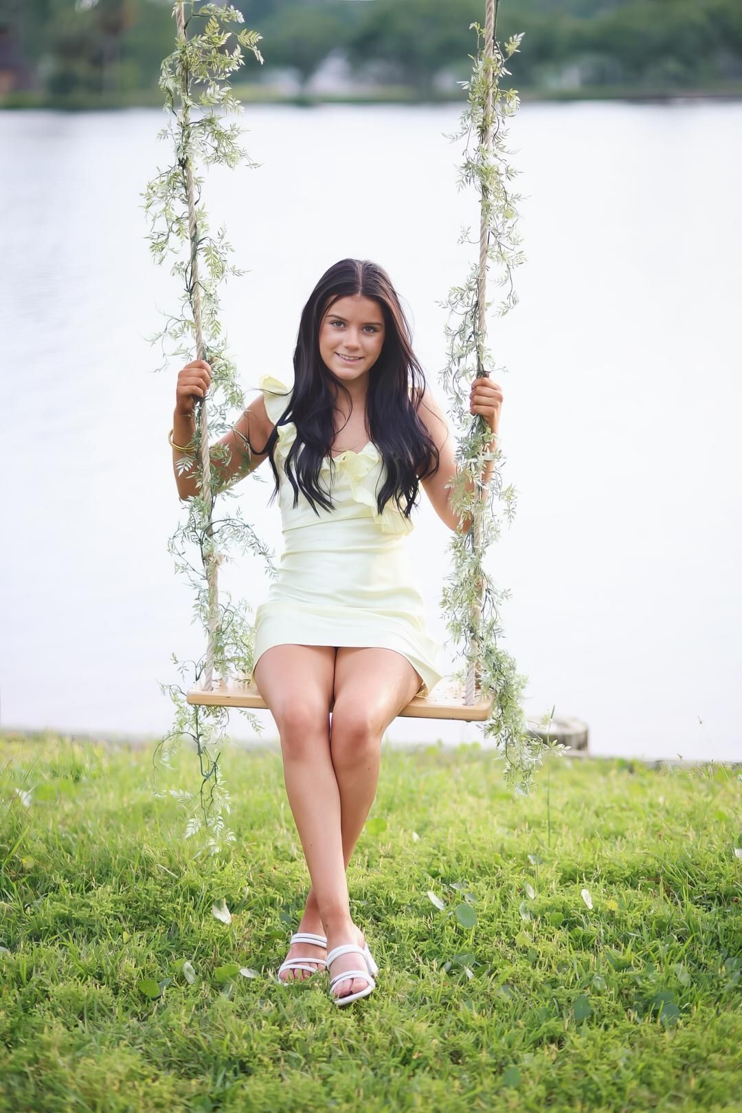 A person in a white dress sits on a rustic wooden swing by a lake surrounded by green grass.