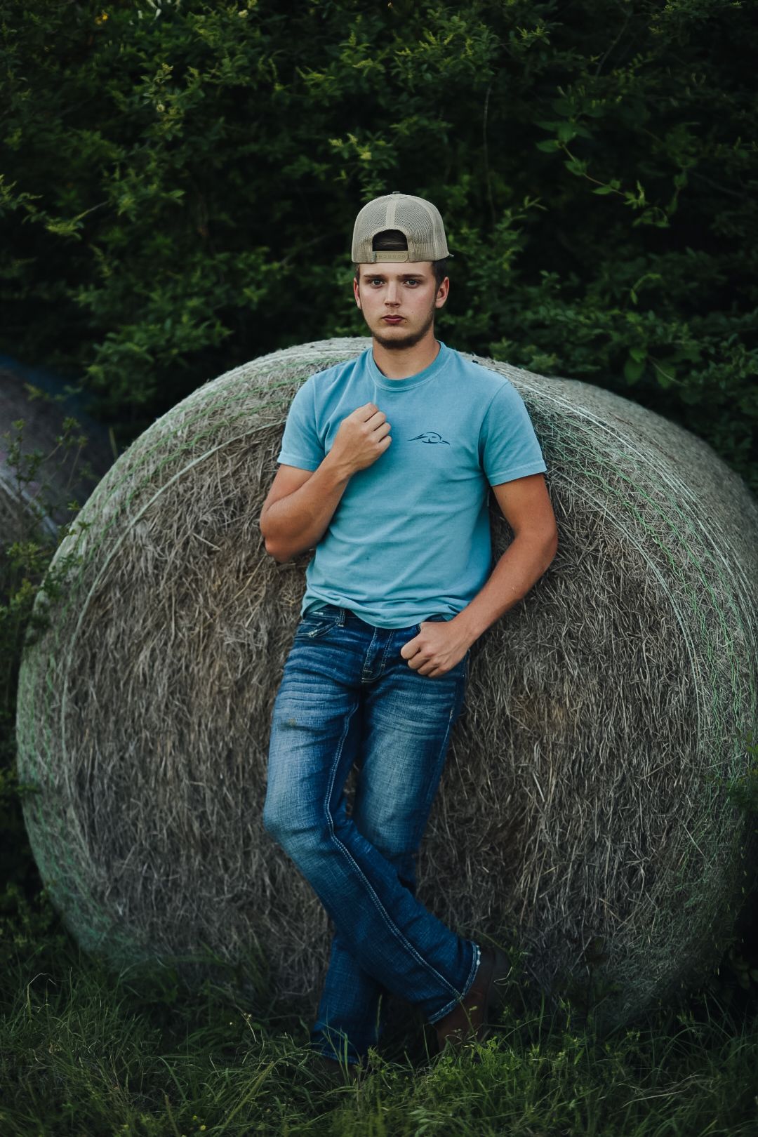 Person in blue t-shirt and jeans sits casually on large round hay bale in outdoor rural setting during golden hour.