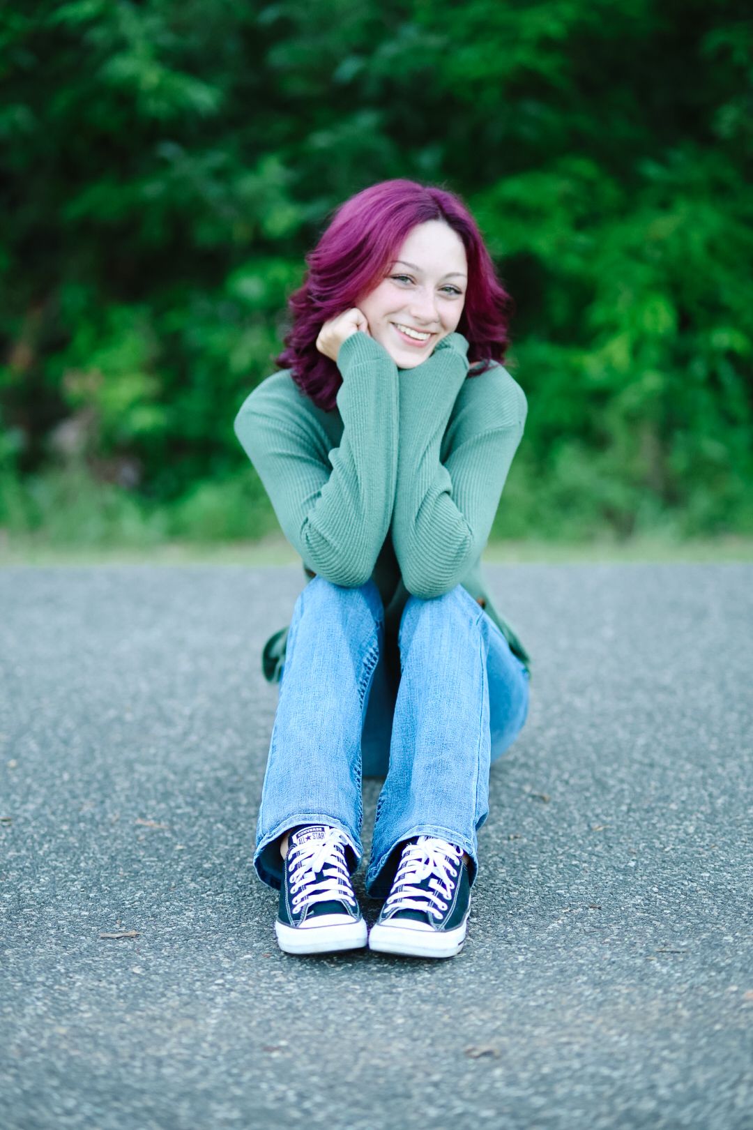 Person in green sweater and jeans sits casually on asphalt ground wearing blue Converse sneakers.