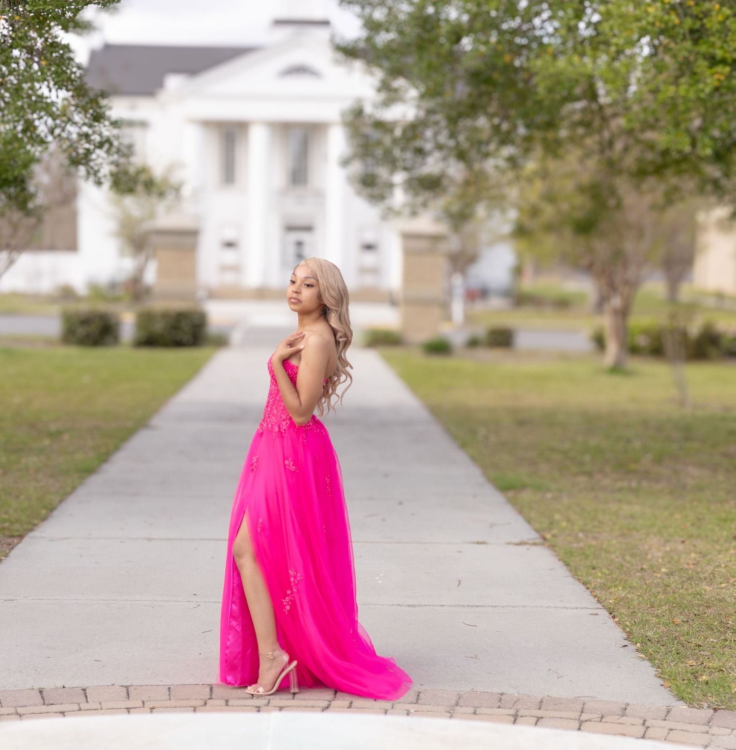 A bright hot pink formal gown with a flowing skirt stands out against the backdrop of a stately white building and pathway.