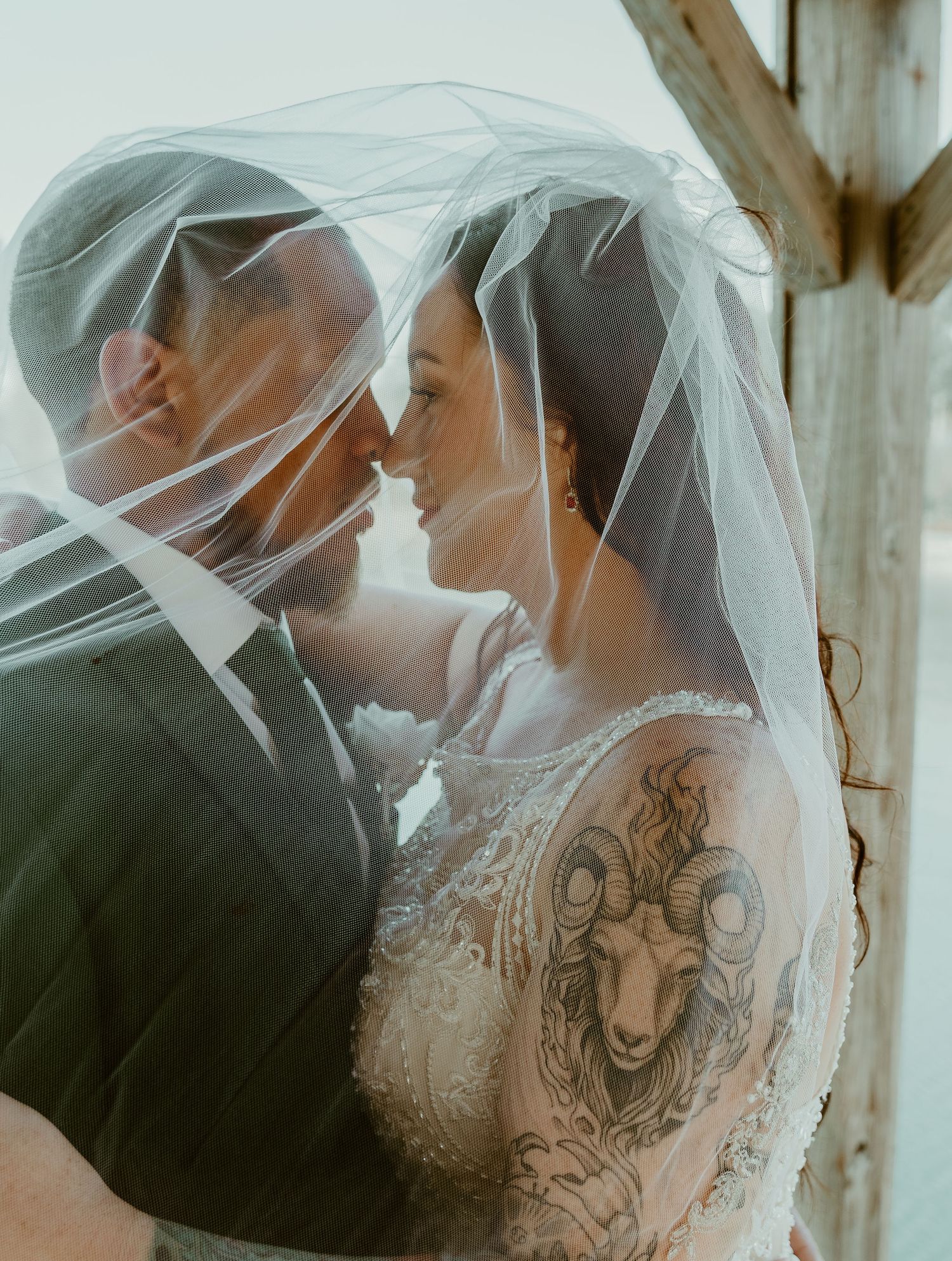 A romantic wedding couple shares an intimate moment under a flowing white veil in a rustic setting.