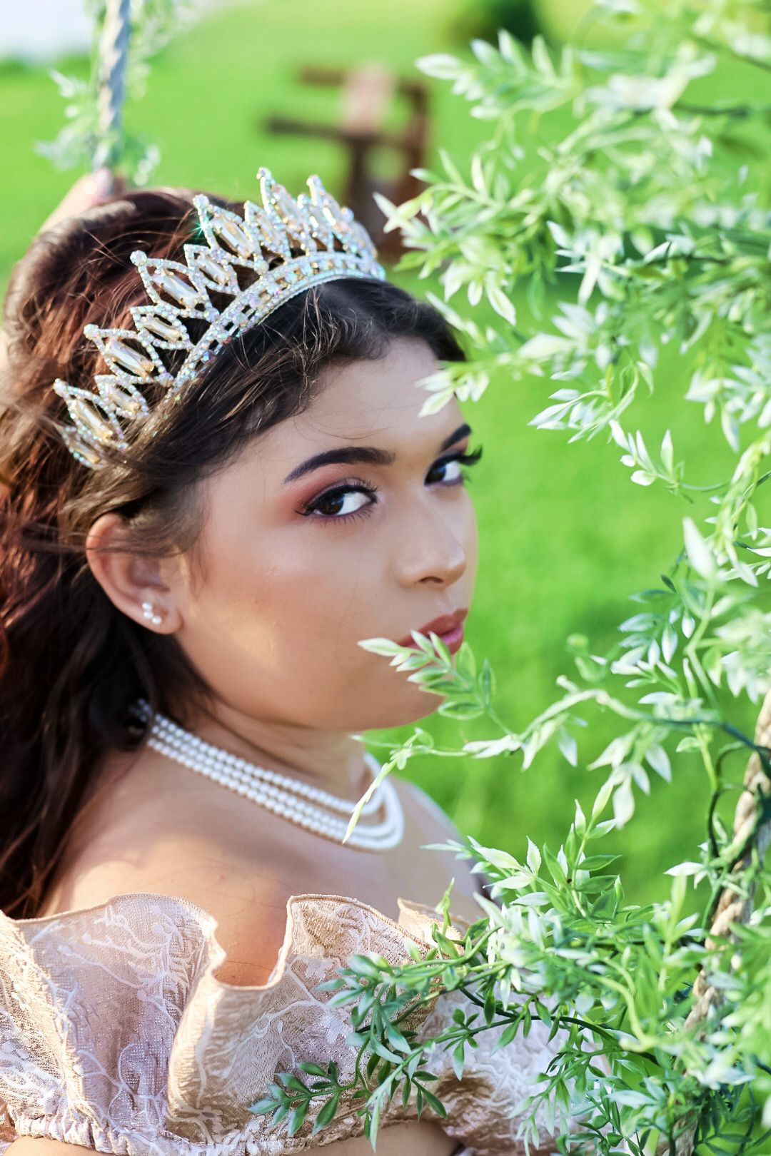 Young woman wearing a crystal tiara, pearl necklace, and elegant gown poses among green leaves outdoors.