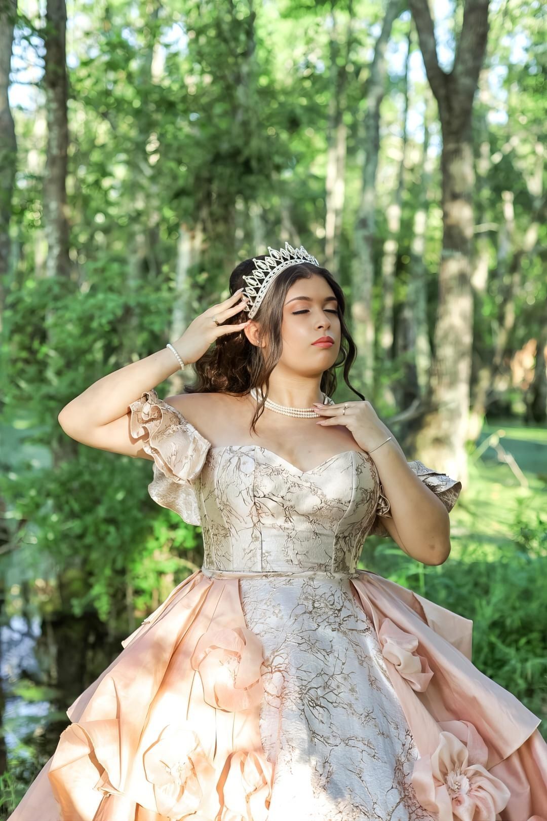 Young woman in elegant blush ballgown and tiara poses gracefully in a lush green forest setting.