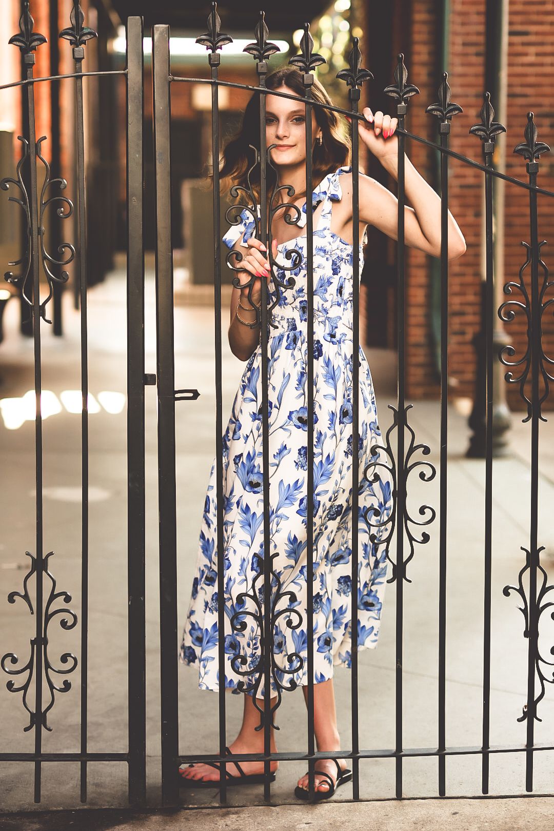 Young woman in blue floral midi dress stands at ornate iron gate in urban setting.