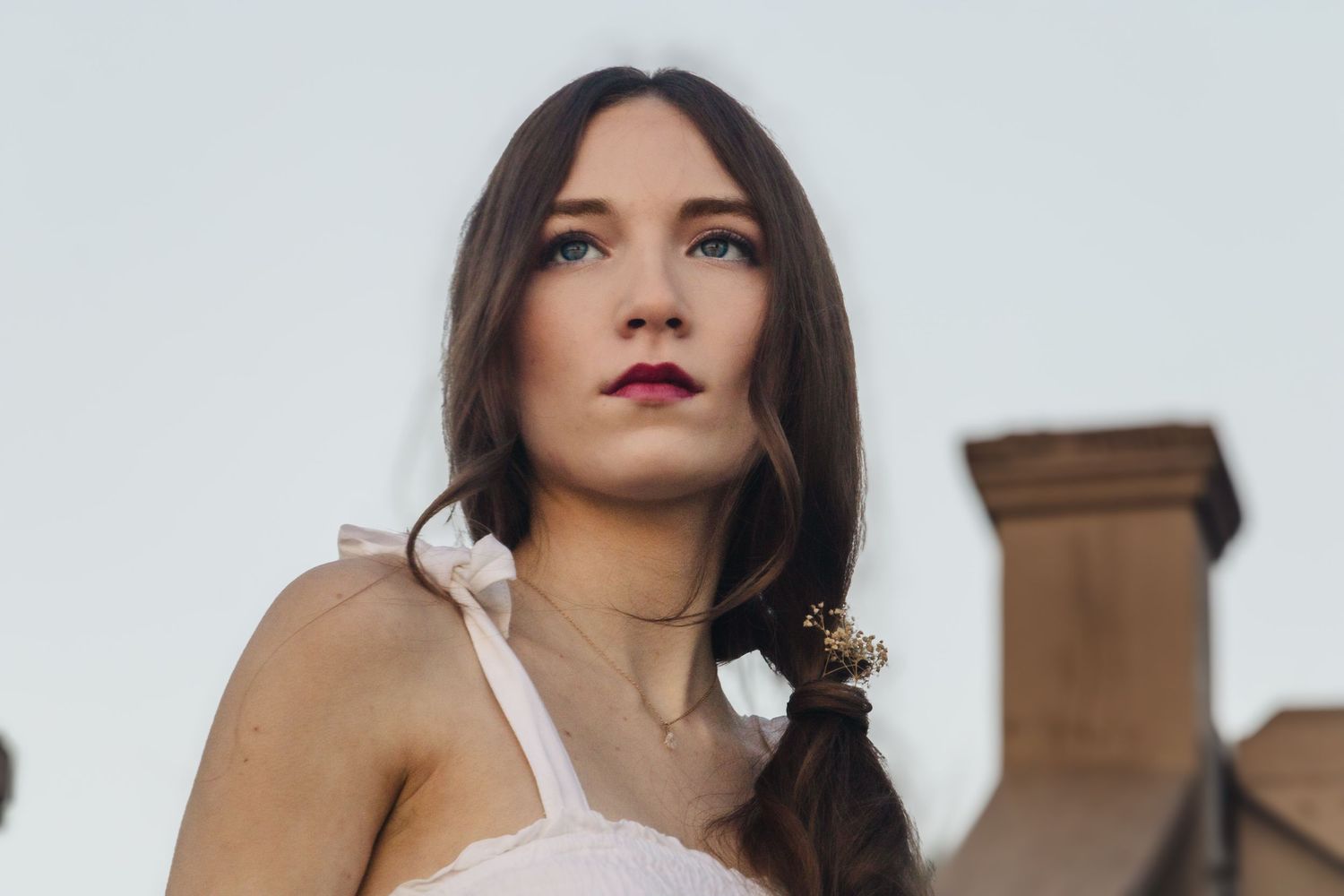 Young brunette woman in white dress gazes upward outdoors, with rooftops and sky in the background.