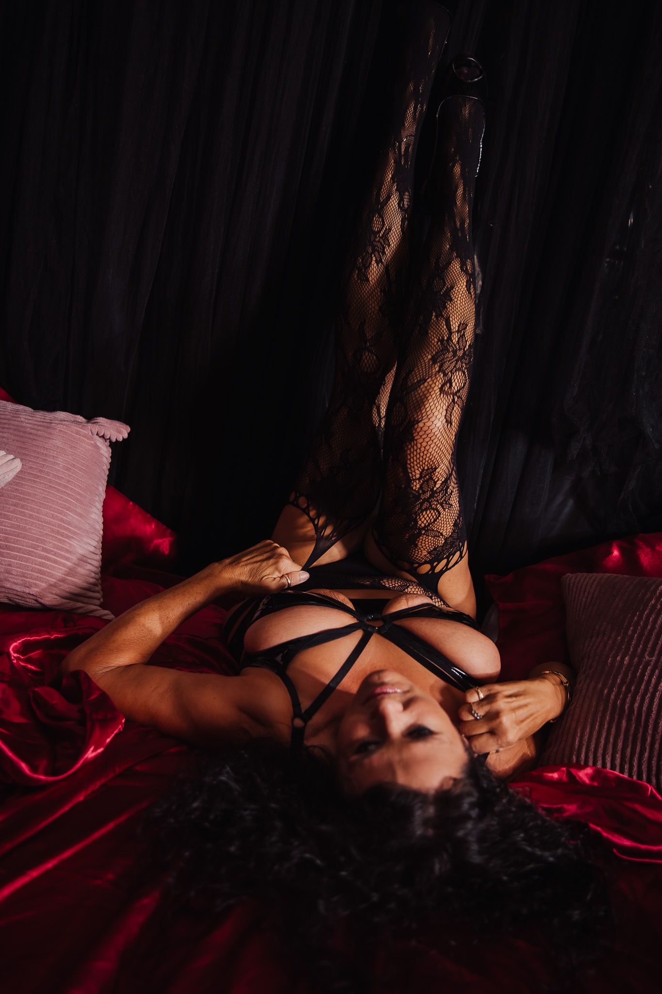 Woman in black lace lingerie and stockings posing on red satin sheets in dramatic dark lighting.