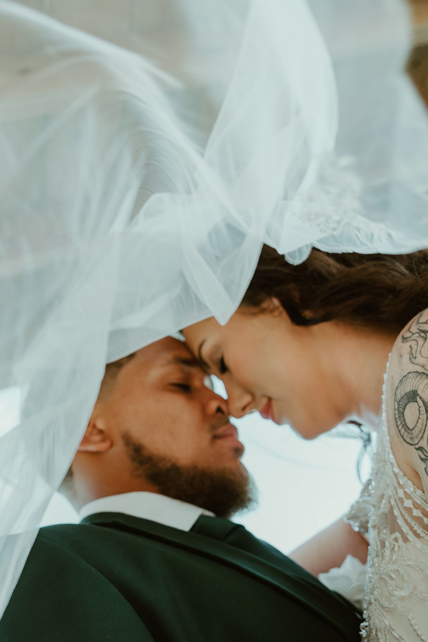 A bride and groom share an intimate forehead touch under a flowing white veil on their wedding day.