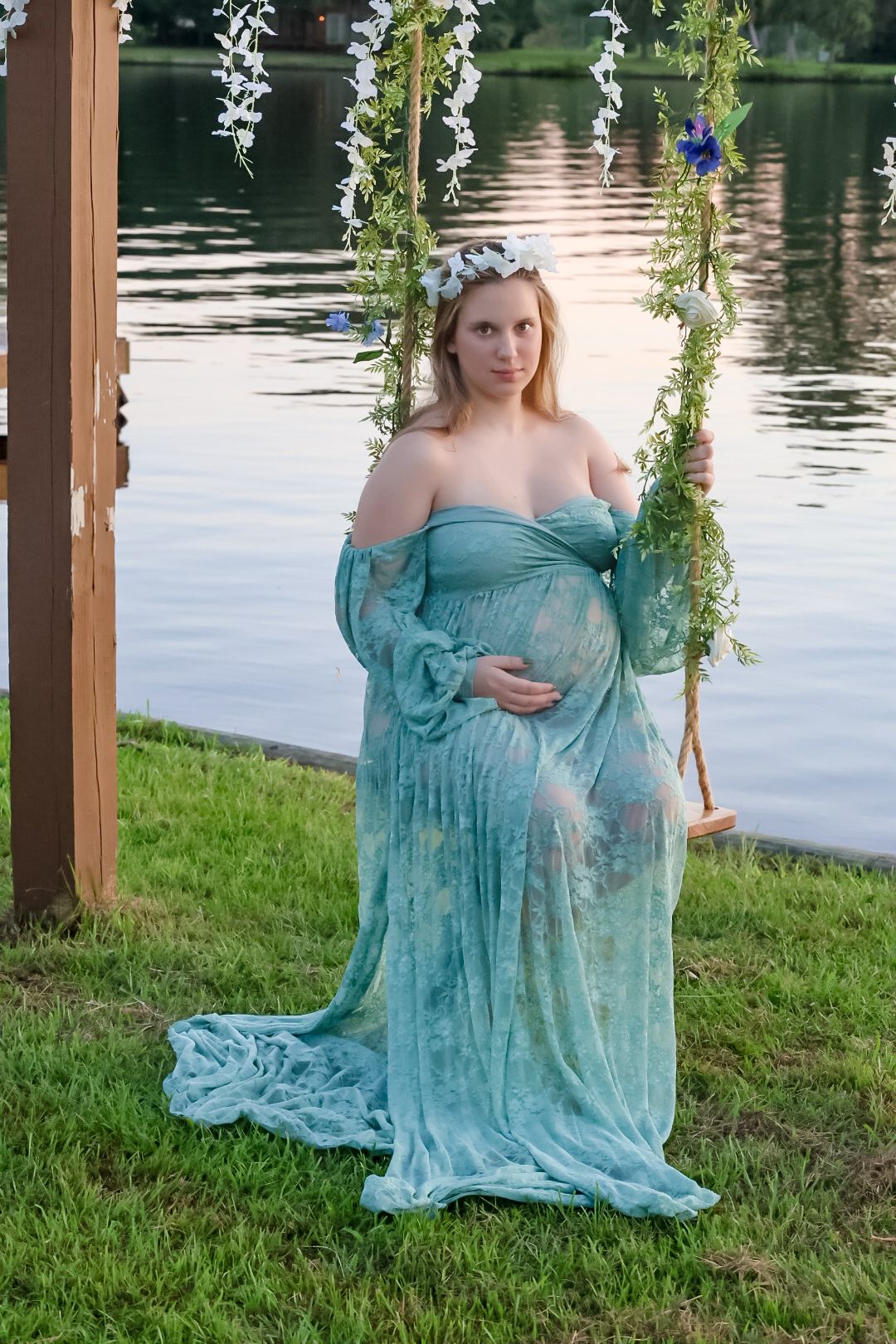 A serene maternity photoshoot by a lake features a turquoise lace dress and hanging floral vines in a wooden archway.