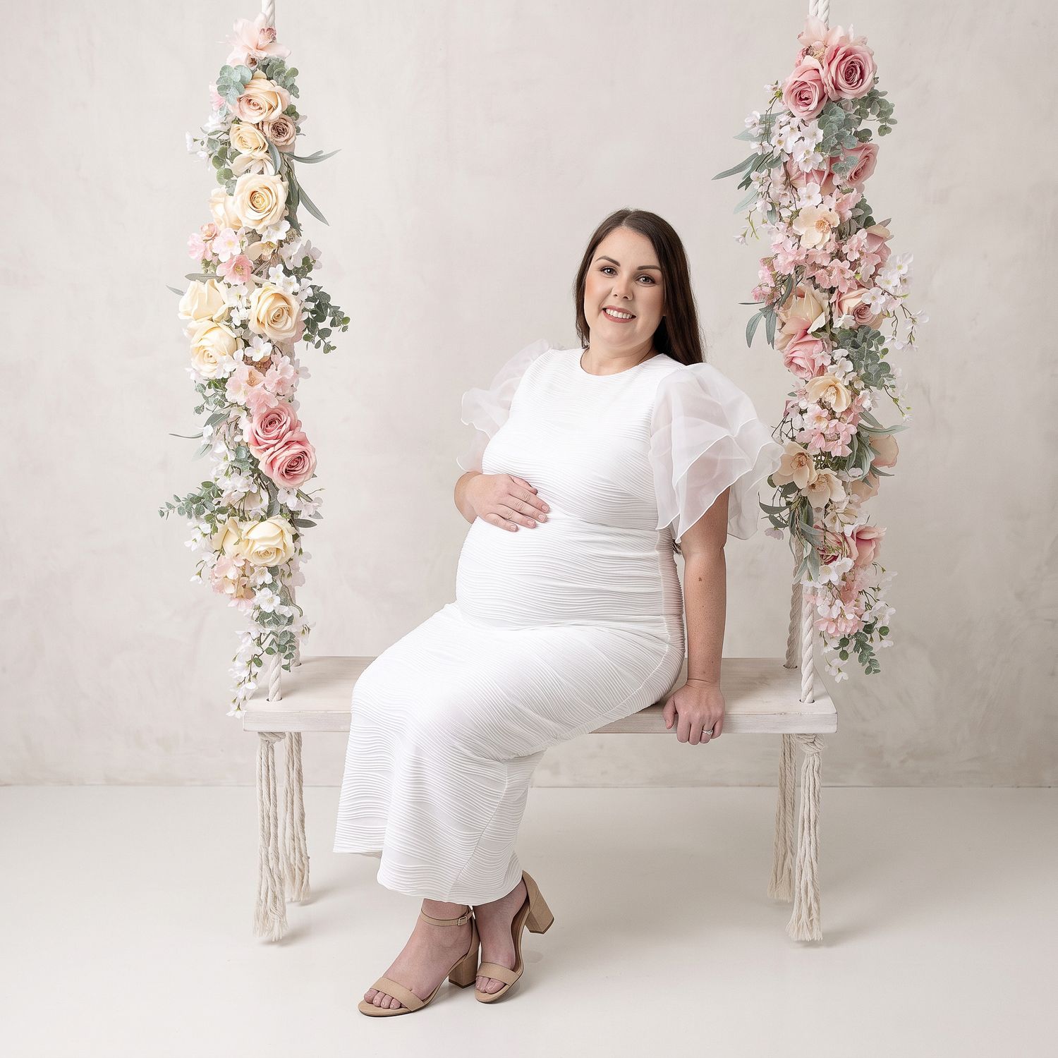 Pregnant woman in white dress sitting on floral swing for maternity photo session.