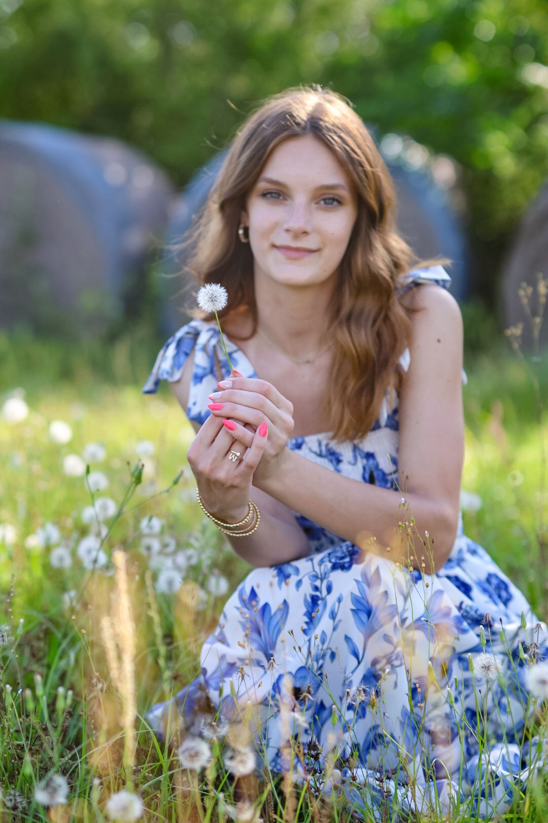 Young woman in blue floral dress sits in meadow holding dandelion with pink manicure.