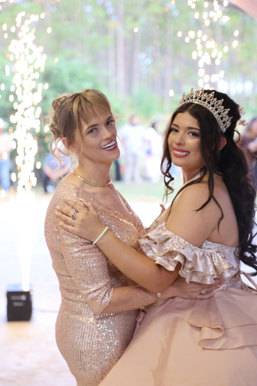 Two women in elegant sequined gowns embrace and smile at a formal celebration with twinkling lights in background.
