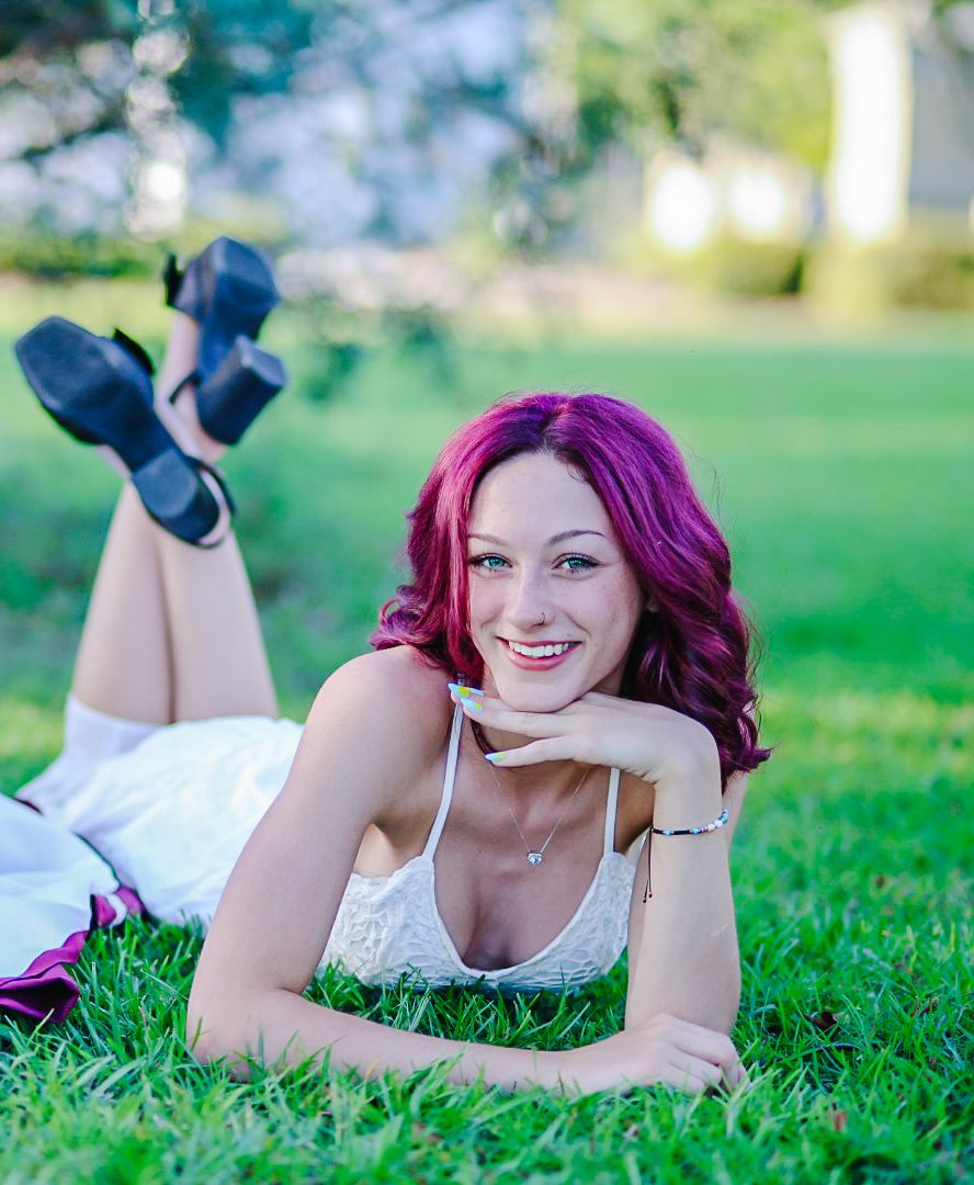 A person with vibrant magenta hair lies on green grass wearing a white dress.