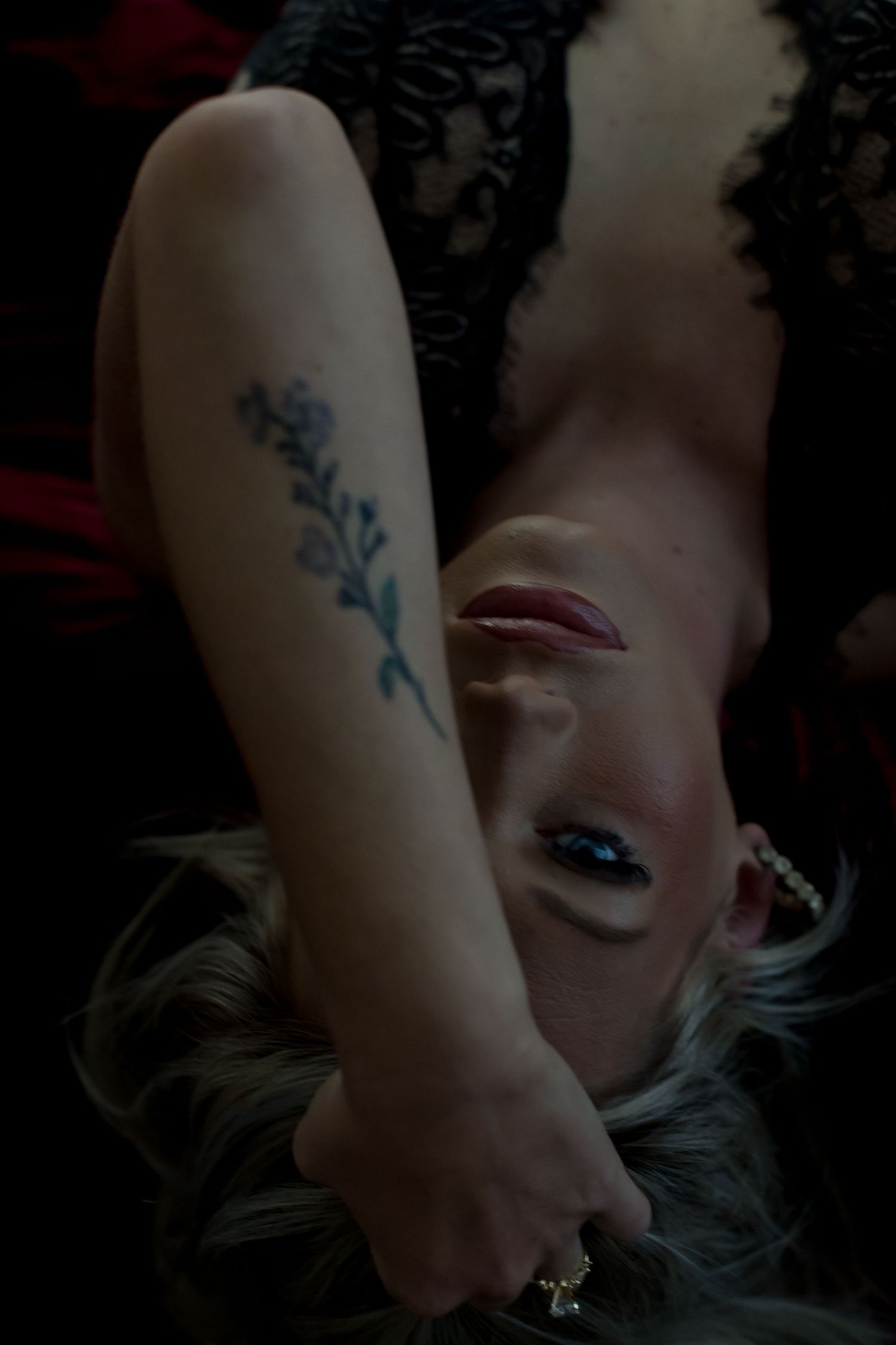 Woman lying down with floral tattoo on forearm, dramatic dark makeup, blonde hair, wearing black lace, moody lighting.