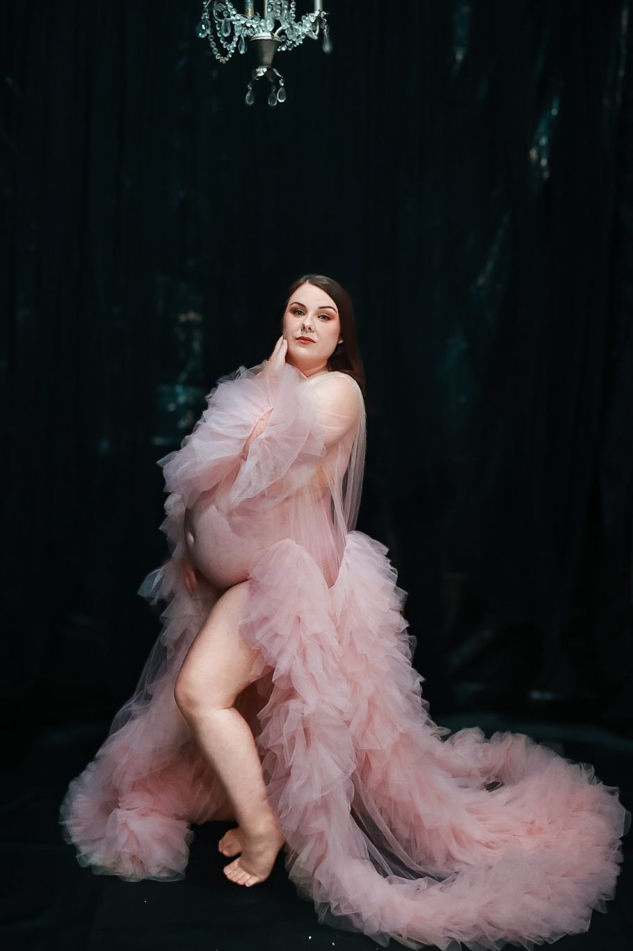 Pregnant woman in sheer pink ruffled maternity gown posing elegantly in dark studio with crystal chandelier.