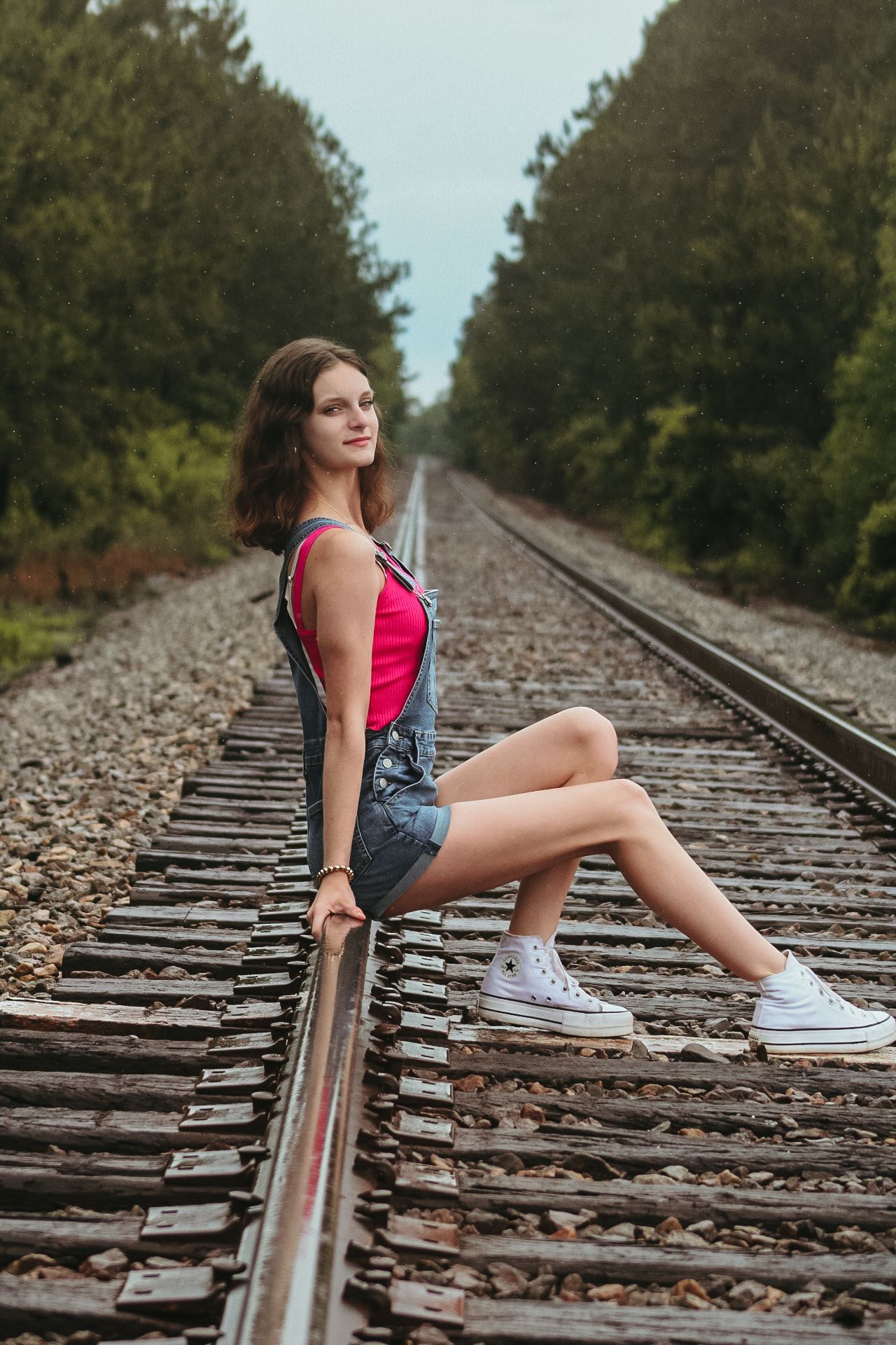 Young woman in denim overalls and pink top posing on railroad tracks surrounded by lush green trees.