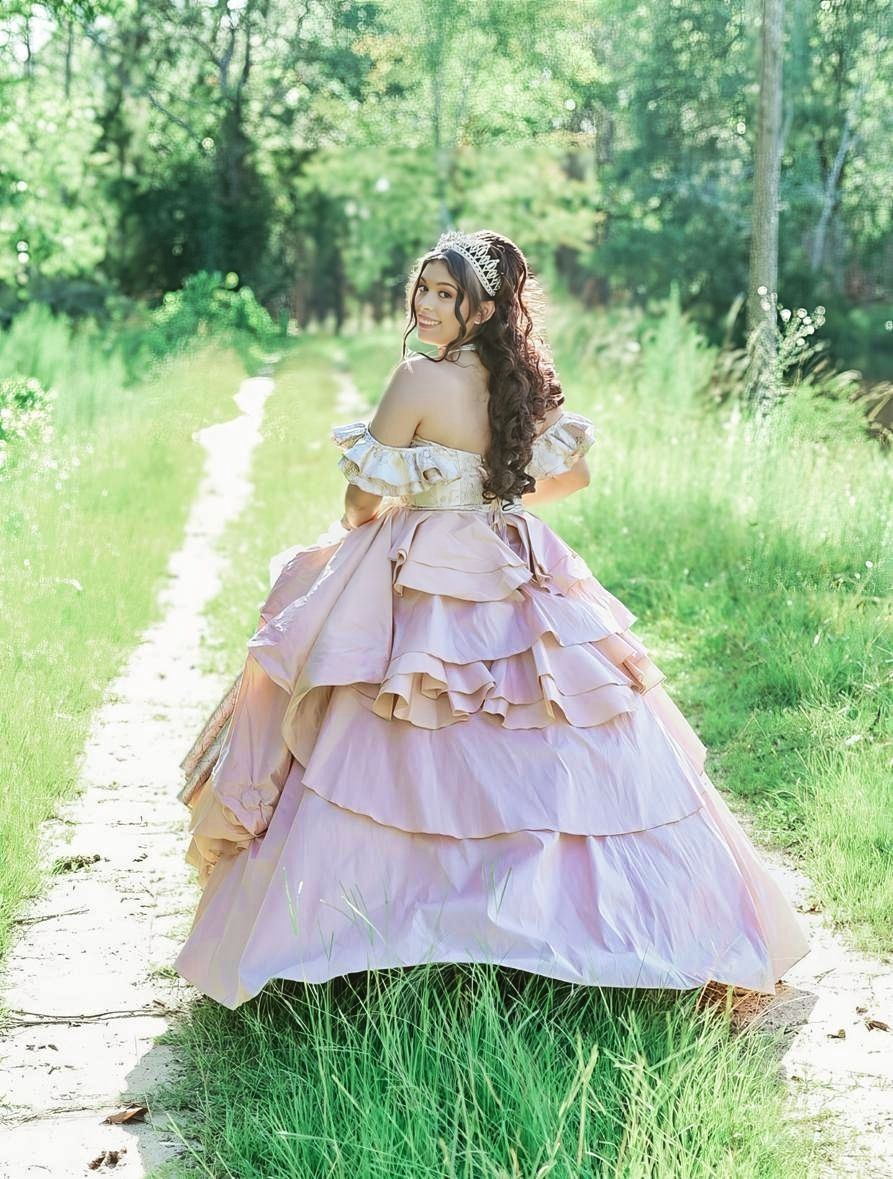 Young woman in a layered blush pink ball gown with off-shoulder design and tiara poses on a garden path.