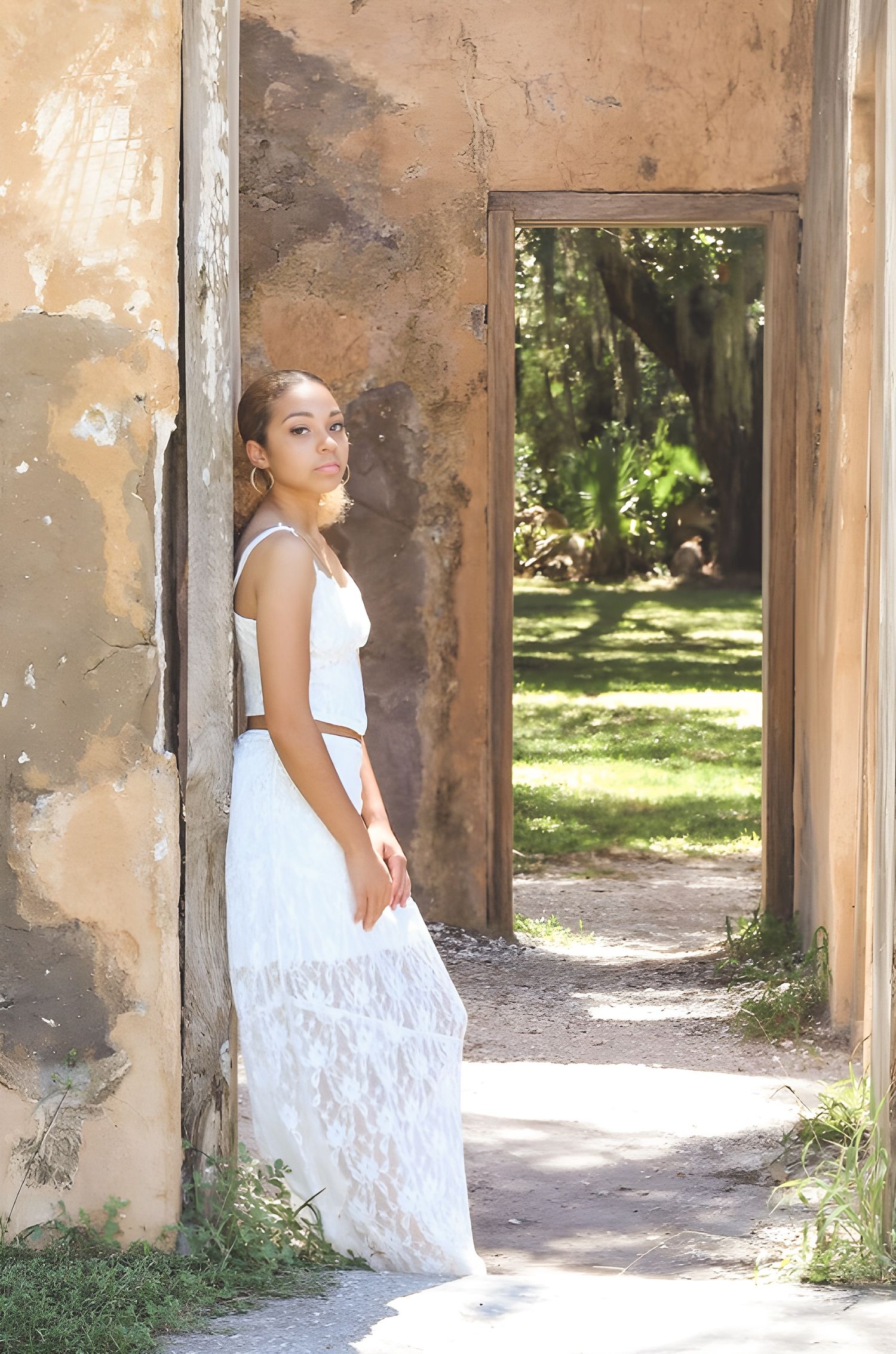 A woman in a flowing white lace dress poses thoughtfully against weathered stone columns in a rustic outdoor setting.