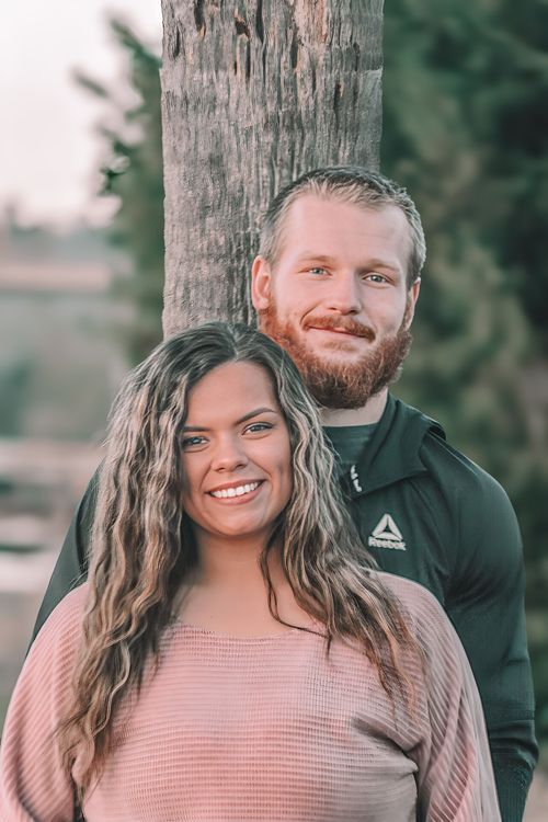 A smiling couple poses outdoors near a tree, woman in pink sweater and man in dark Reebok jacket with red beard.