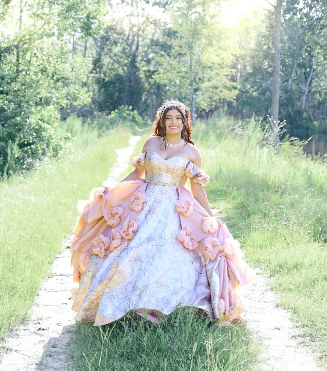 Young woman in ornate pink and gold ball gown with floral details poses on a garden path surrounded by lush greenery.