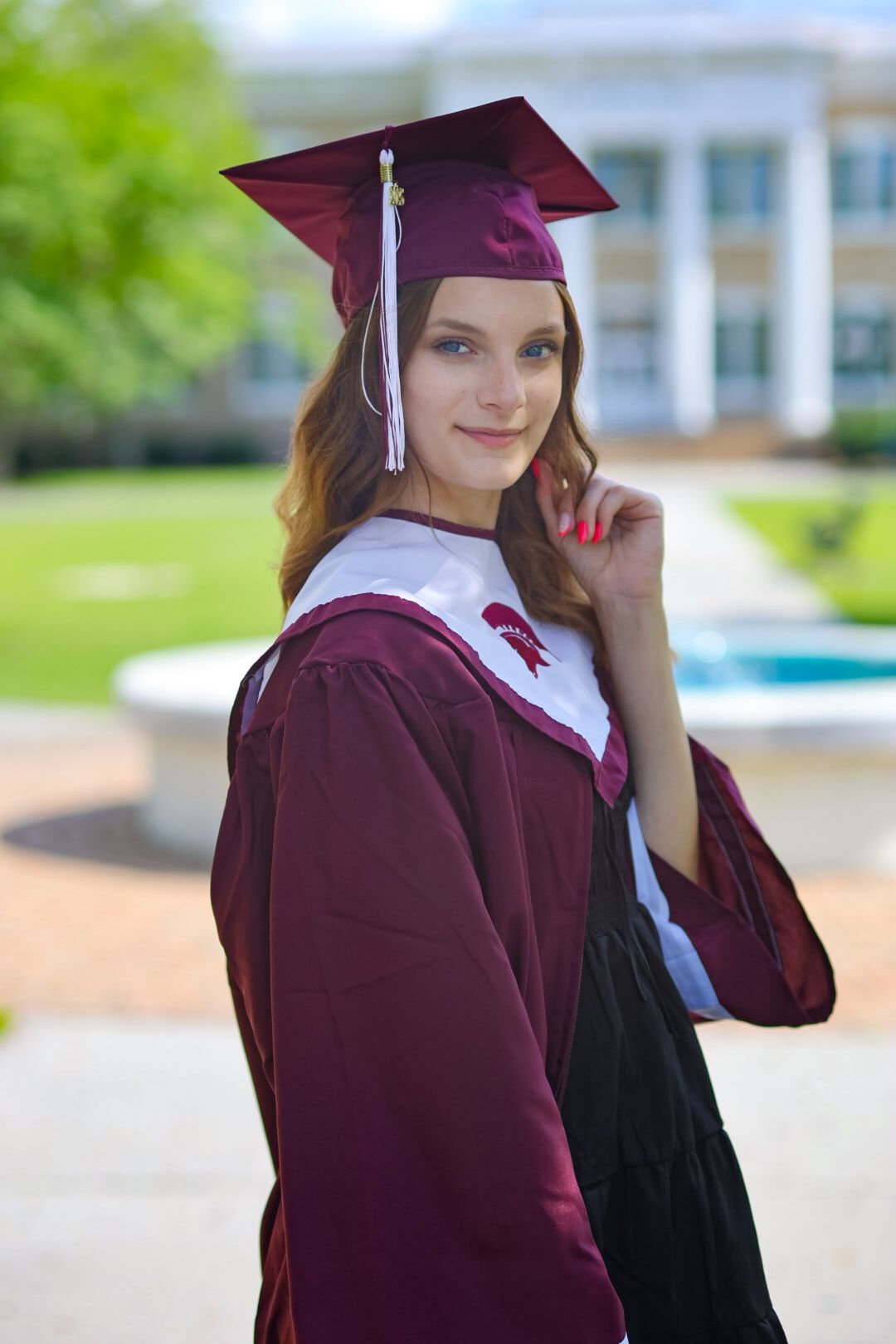 Young woman in maroon graduation cap and gown smiling outdoors near a fountain on a college campus.