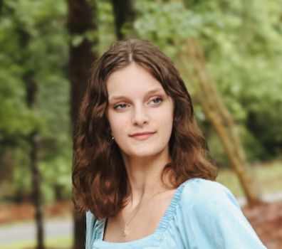 Young woman with brown wavy hair smiling outdoors in a light blue top, surrounded by lush green trees.