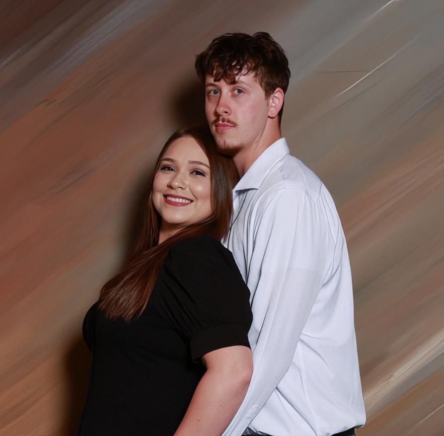 A young couple posing together, woman in black dress and man in white shirt, against a warm blurred background.