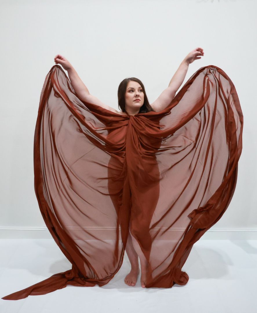 Woman in flowing rust-red chiffon gown poses gracefully with arms raised, fabric spread like wings against white background.