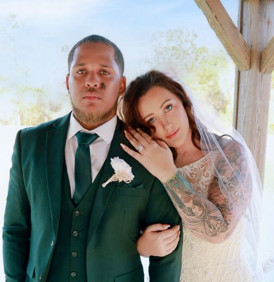 Newlywed couple in formal wedding attire pose together under a rustic wooden structure outdoors.