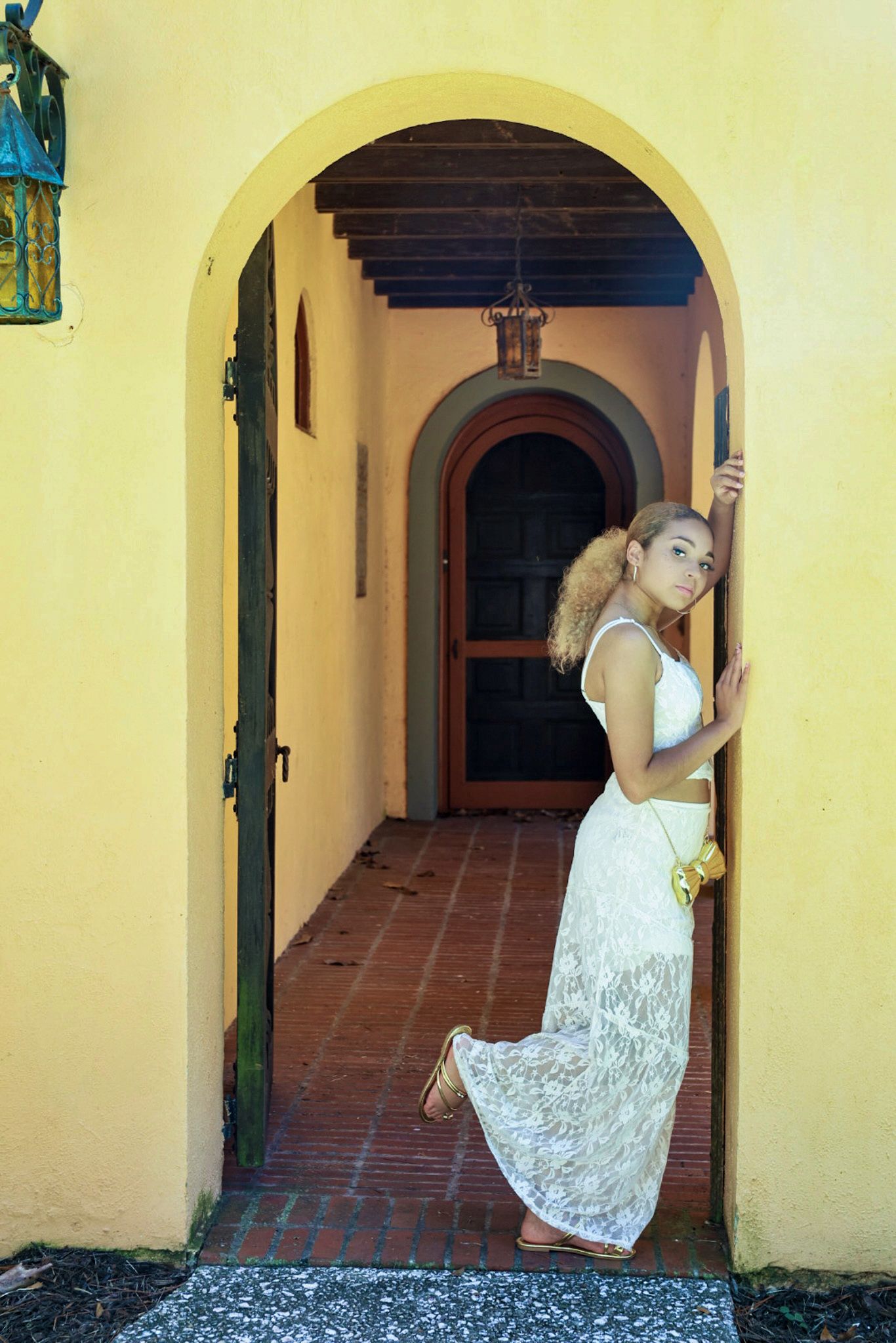 A woman in a white lace dress poses in an arched doorway of a yellow Mediterranean-style building.