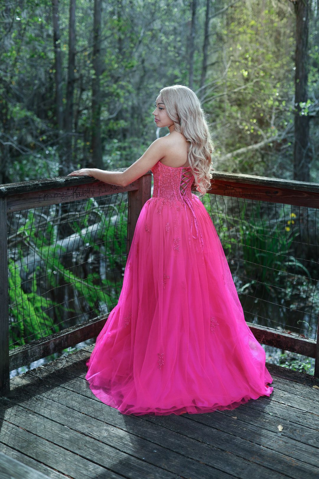 A person in a flowing hot pink ballgown twirls on a wooden deck surrounded by lush forest greenery.
