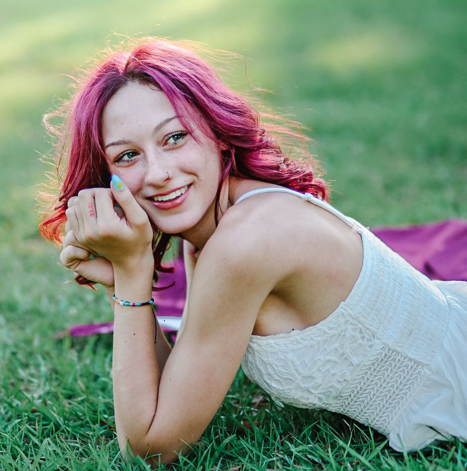 Figure with magenta hair in white lace top relaxes on grass in summer sunlight.