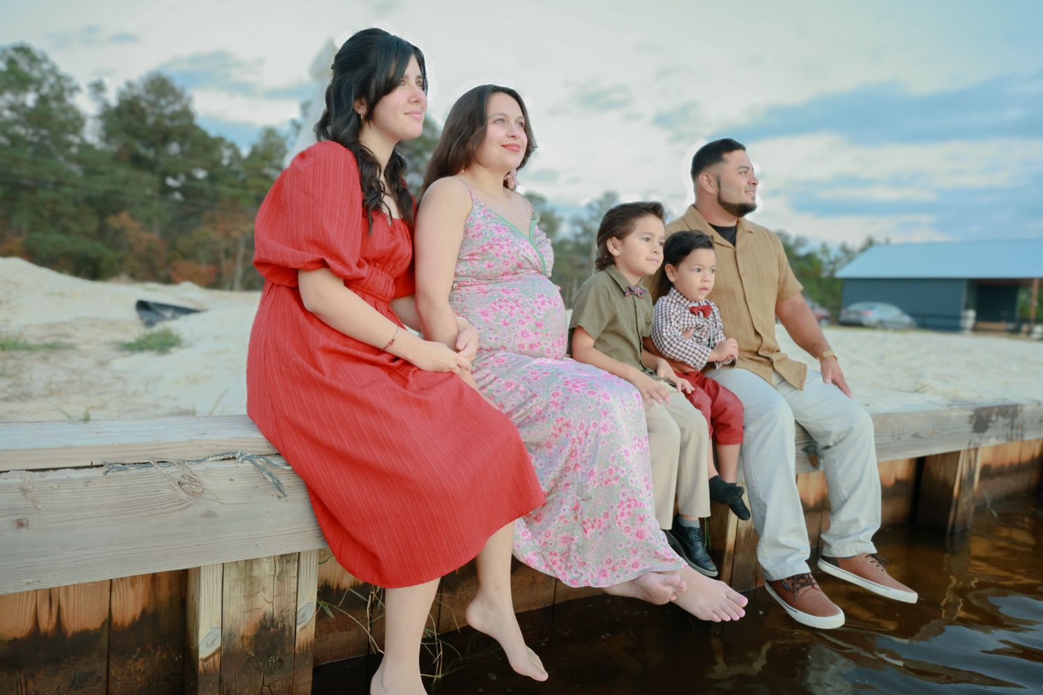 A family sits together on a wooden dock by the water during sunset, wearing casual dresses and clothing.
