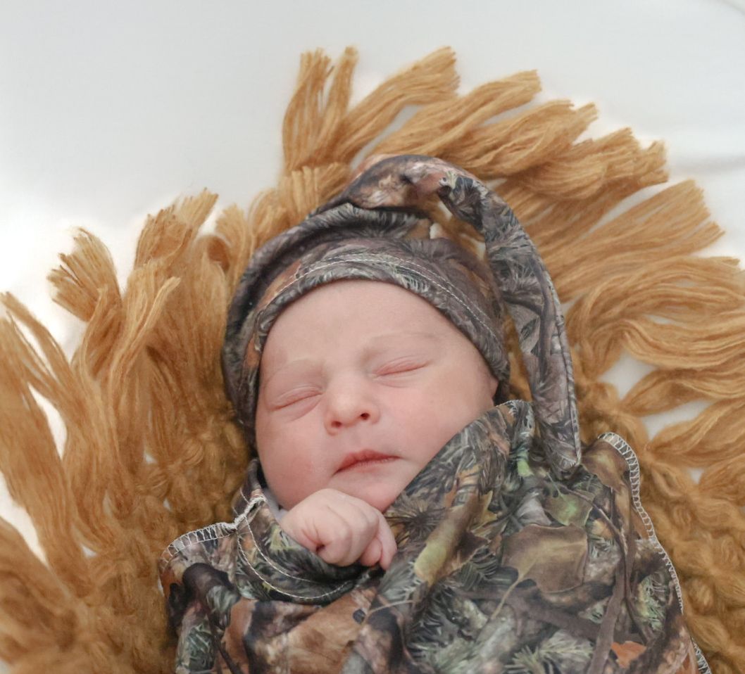 A sleeping baby wearing camouflage clothing lies on soft blonde fur with eyes closed peacefully.