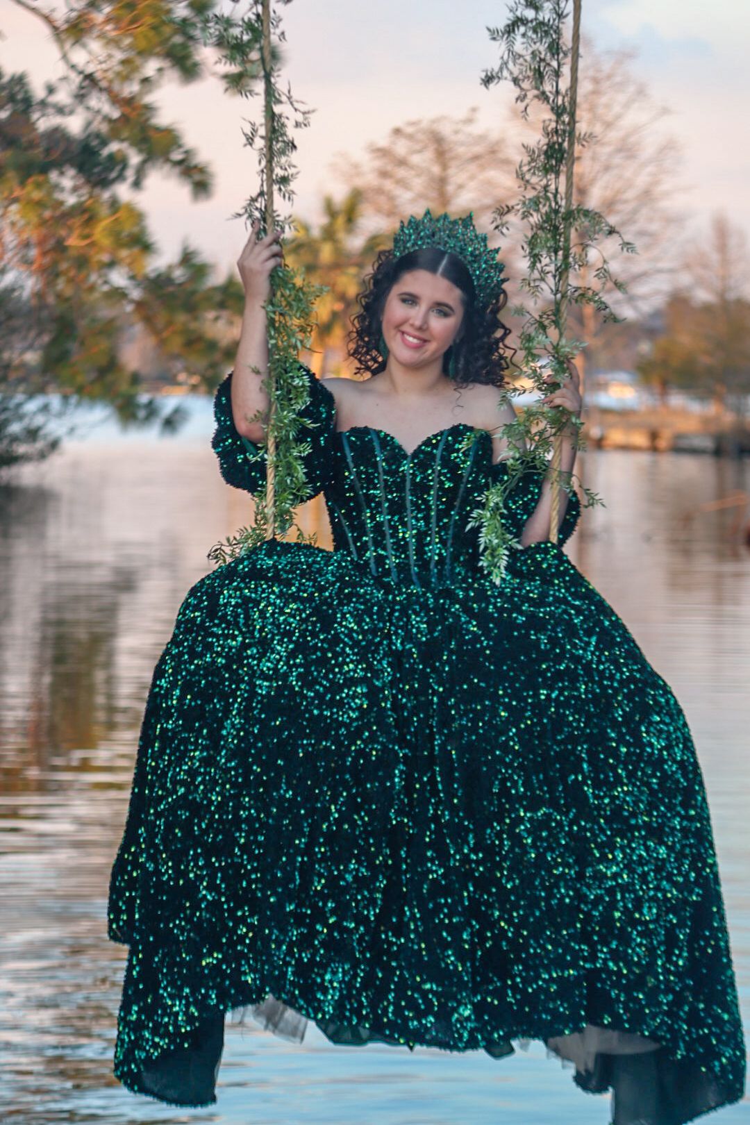 A person in a sparkling emerald green gown poses by a waterfront at sunset with trees in the background.