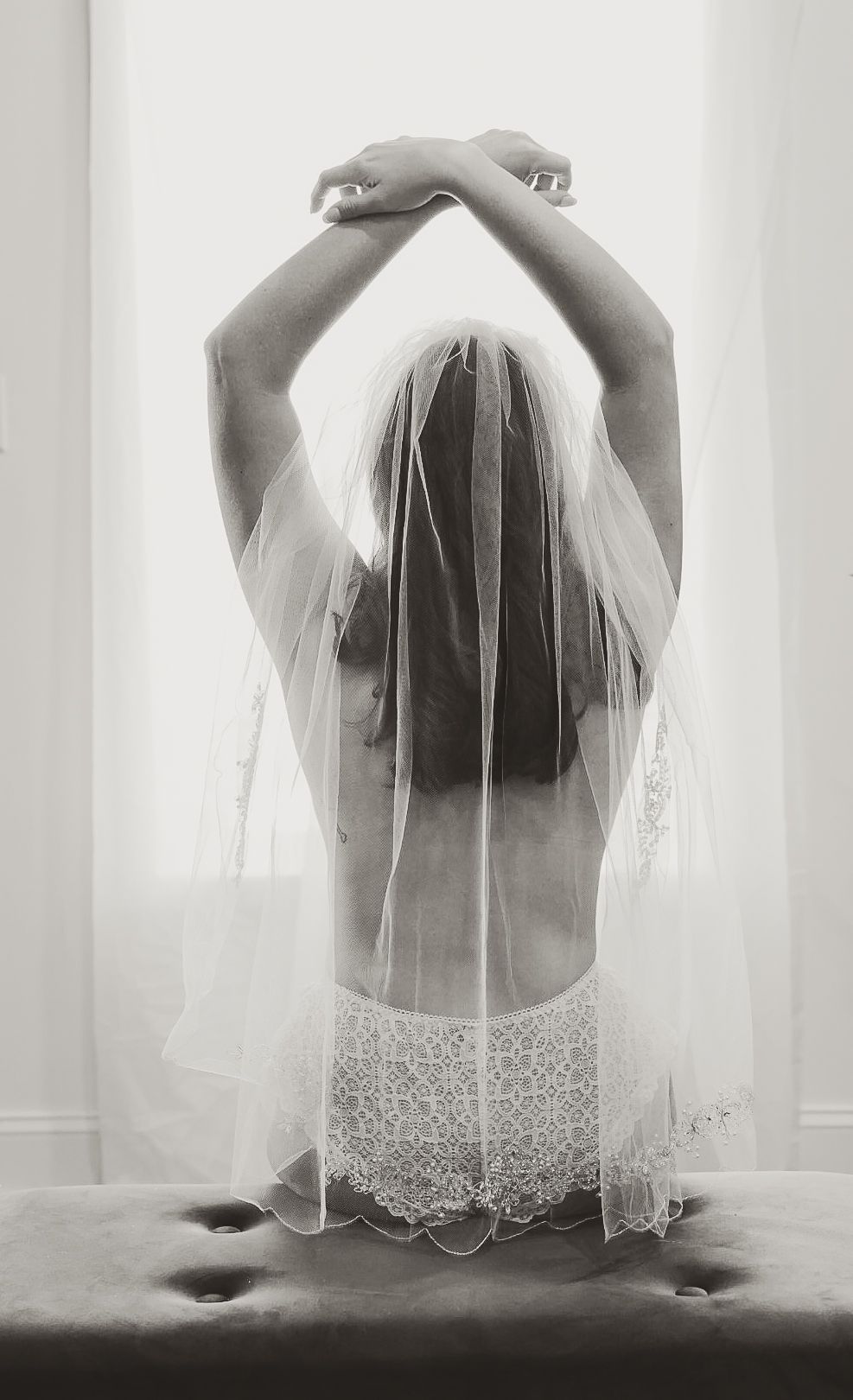 Black and white photo of a bride in lace lingerie and veil, arms raised gracefully, viewed from behind near a window.