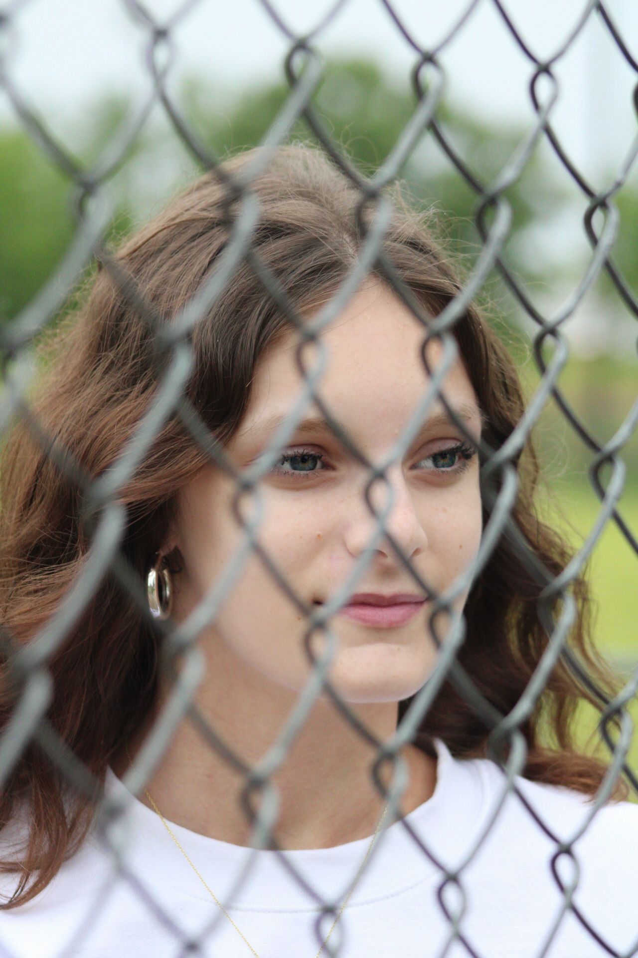 Young woman with brown hair gazes through a chain-link fence outdoors, wearing a white top and hoop earrings.