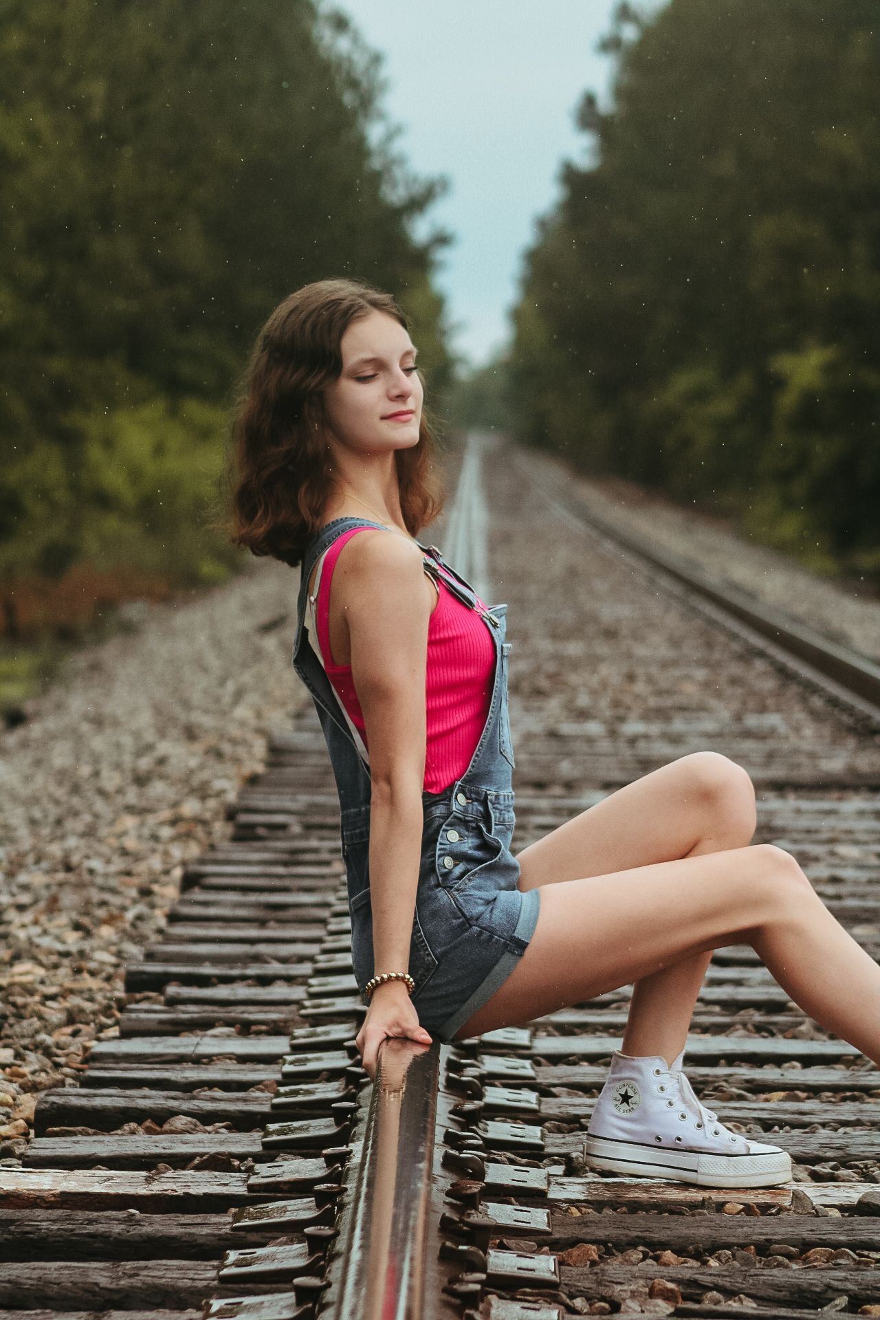 Young woman in denim overalls and pink top poses on railroad tracks surrounded by lush green trees.