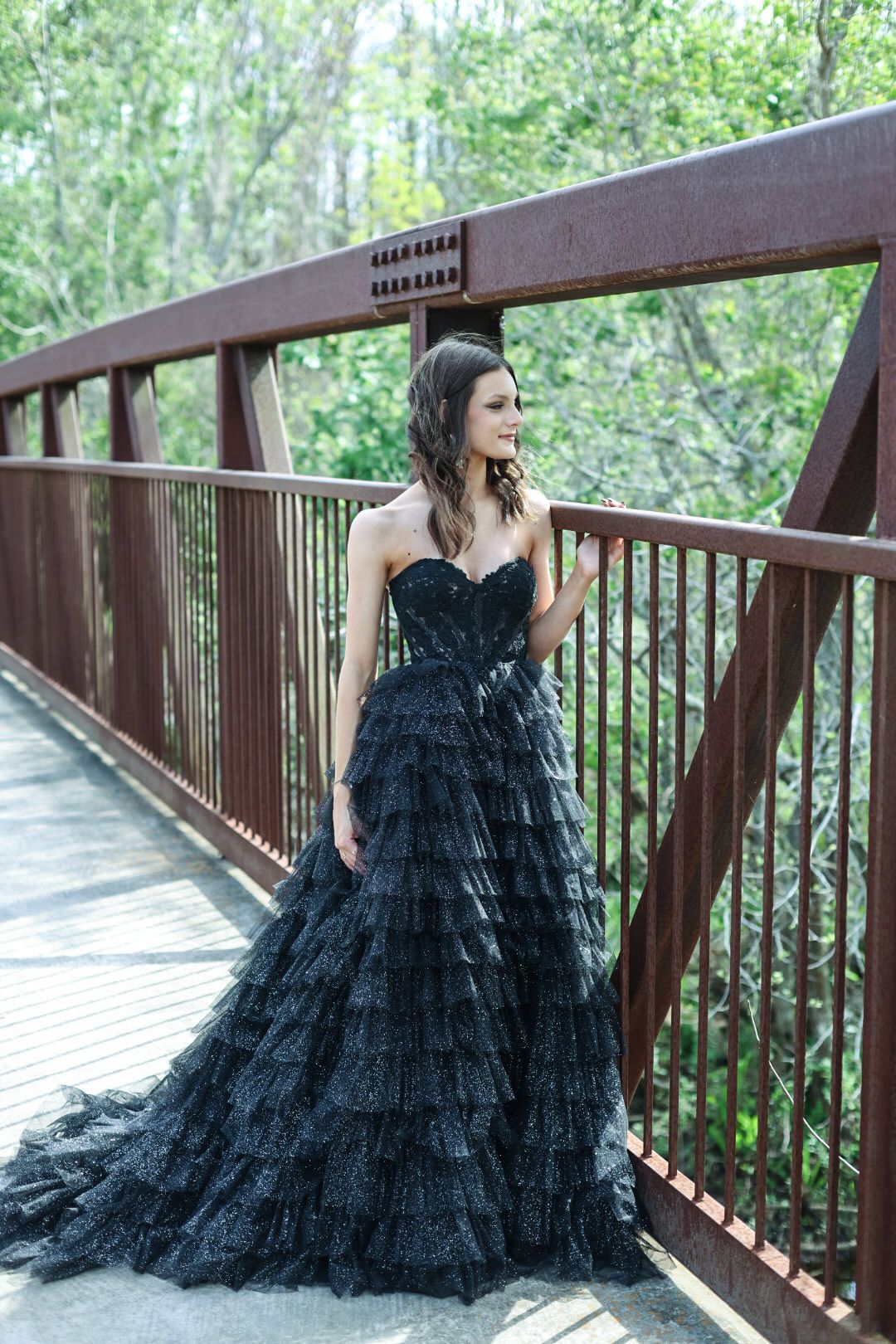 Woman in black strapless ruffled ball gown posing on a rustic wooden bridge surrounded by lush greenery.